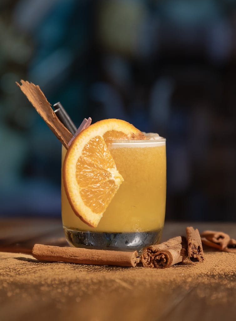 Close-up of a citrus cocktail garnished with an orange slice and cinnamon stick on a wooden table.