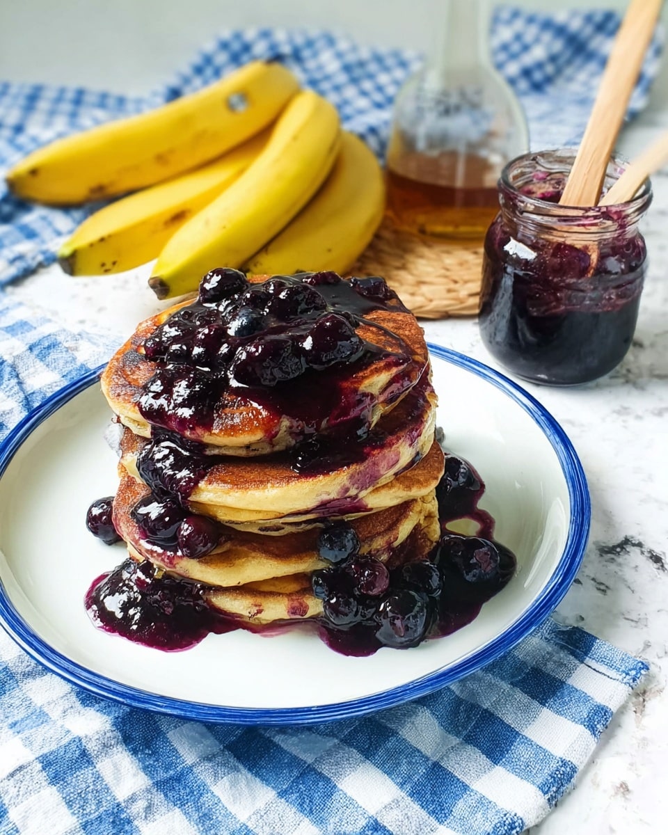 A stack of four golden-brown pancakes sits on a white plate with a blue rim, generously covered with dark purple blueberry sauce that drips down the sides and pools on the plate. Small whole blueberries are scattered on top and around the pancakes, adding texture and color contrast. Behind the plate, three yellow bananas rest on a white marbled surface covered partly by a blue and white checkered cloth. Next to the bananas, there is an open jar of dark blueberry jam with a wooden spoon inside and a clear bottle with amber liquid. The scene is bright with natural light. photo taken with an iphone --ar 4:5 --v 7