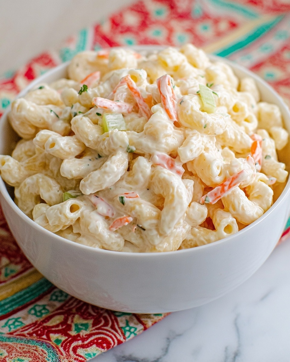 A white bowl is filled with creamy macaroni salad consisting of three visible layers: the main layer of soft, pale yellow macaroni pasta coated in a thick white mayonnaise dressing, mixed throughout with thin strands of light orange shredded carrot and small pieces of pale green celery, plus tiny flecks of darker green herbs sprinkled evenly. The bowl is sitting on a white marbled surface with a colorful cloth with red, green, and patterned designs partly visible beneath. The image looks close-up and bright. Photo taken with an iphone --ar 4:5 --v 7