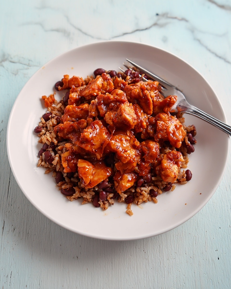 A white plate holds a dish with two layers: the bottom layer is a mix of brown rice and dark red beans, showing a grainy texture and scattered beans, while the top layer is made of chunky, crispy pieces of reddish-brown cooked chicken glazed with a shiny sauce. A metallic fork rests on the right side of the plate, partially touching the food. The plate is set on a white marbled surface. photo taken with an iphone --ar 4:5 --v 7