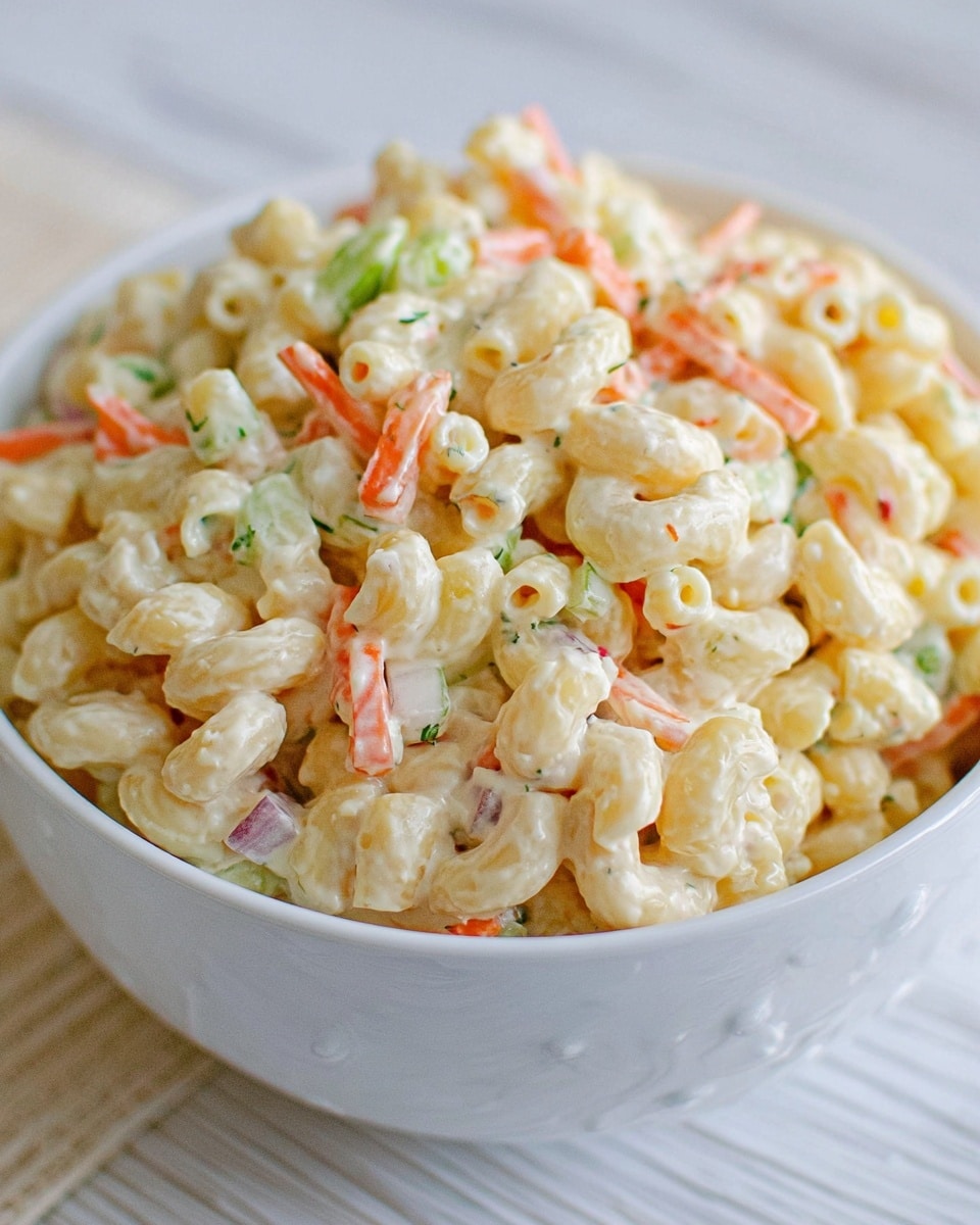 A close-up view of a white bowl filled with creamy macaroni salad placed on a white marbled surface. The salad has three visible layers: the base layer consists of small elbow macaroni pasta coated in a smooth, creamy dressing that is pale yellowish-white in color and glossy in texture. Mixed within the pasta are thin strips of orange carrot and small diced pieces of pale green celery, adding color contrast and slight texture. There are also tiny bits of darker green herbs and small white onion pieces scattered throughout, creating a fresh, mixed appearance. The bowl is full, with the salad slightly mounded over the edge. photo taken with an iphone --ar 4:5 --v 7
