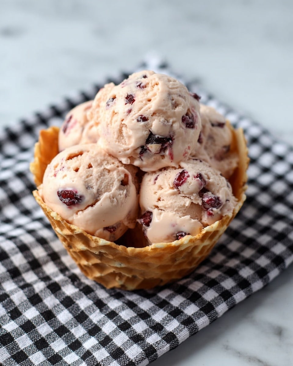 A waffle bowl filled with four scoops of light pink ice cream mixed with dark red berry pieces, placed on a black and white checkered cloth on a white marbled surface. The ice cream scoops have a creamy texture with visible berry chunks spread throughout. The waffle bowl is golden brown with a grid pattern and holds the ice cream neatly. photo taken with an iphone --ar 4:5 --v 7