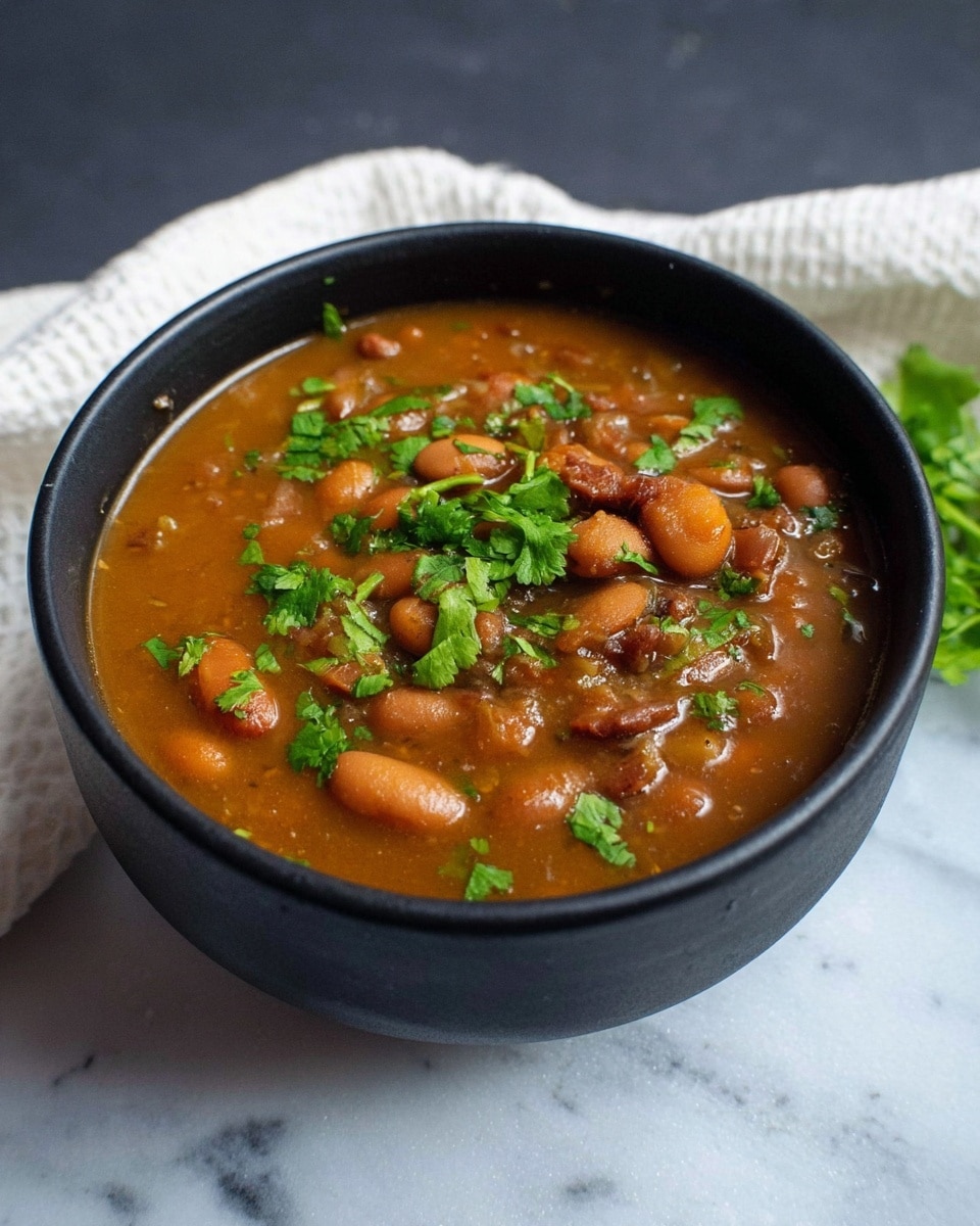 A black bowl filled with brown stew showing many soft beans and small pieces of cooked ingredients in a thick brownish-orange broth, topped with fresh green cilantro leaves scattered on the surface. The bowl sits on a white marbled texture with a white cloth partially visible in the background. photo taken with an iphone --ar 4:5 --v 7