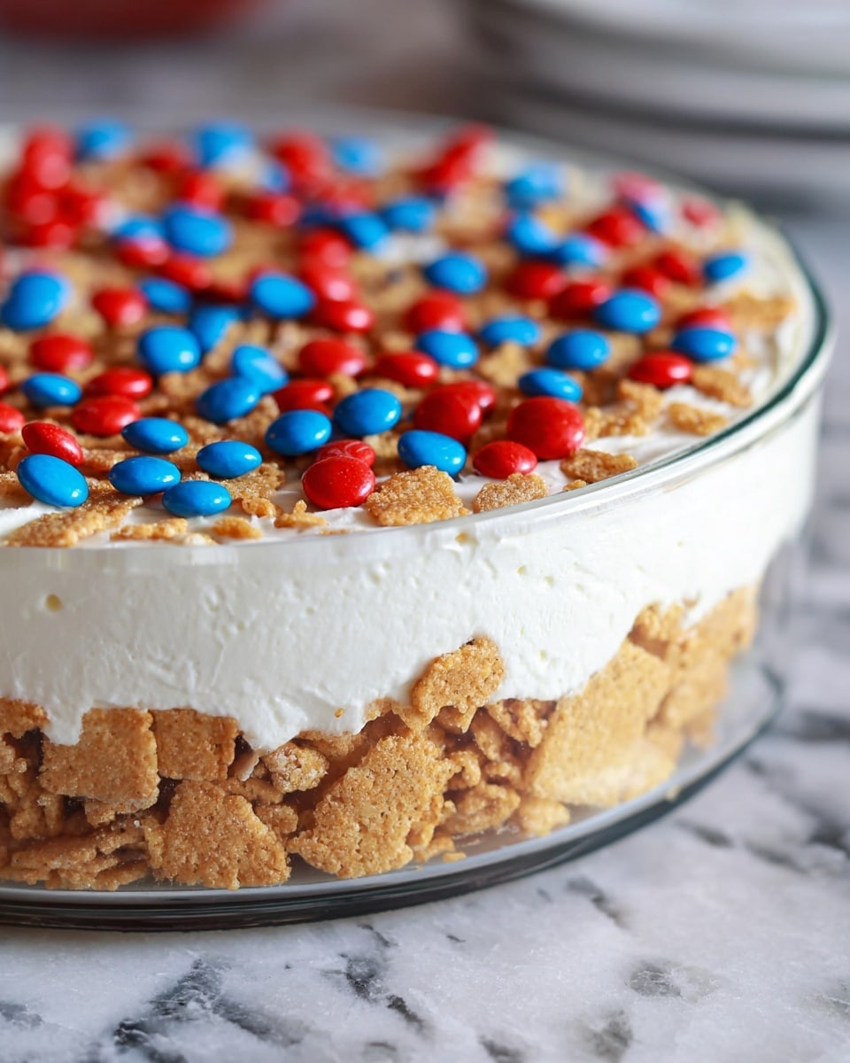 A close-up side view of a two-layer dessert in a clear glass dish, placed on a white marbled surface. The bottom layer is made of broken golden cereal pieces, giving a crunchy texture and light brown color. On top of this is a thick, smooth, white cream layer, spread evenly with a slightly wavy surface. The top of the cream is decorated with scattered small red and blue candy-coated chocolates. Photo taken with an iphone --ar 4:5 --v 7