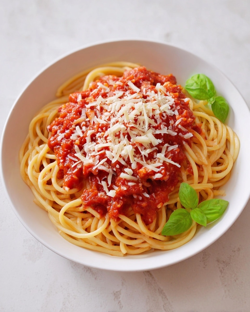 A white shallow bowl holds a serving of spaghetti with tomato sauce, showing about two layers; the bottom layer is the pale yellow pasta, thick and slightly curved, while the top layer is a bright red chunky tomato sauce spread unevenly over the noodles, sprinkled generously with shredded pale yellow cheese. Two small fresh green basil leaves sit on the right side of the bowl near the pasta. The bowl is placed on a white marbled surface. photo taken with an iphone --ar 4:5 --v 7