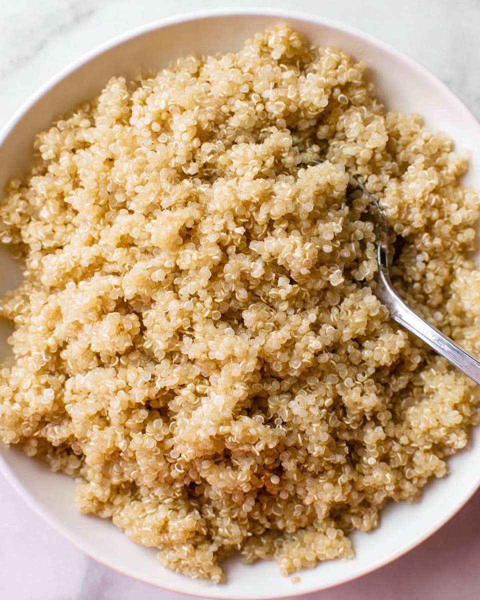 A close-up image of a white bowl filled with cooked quinoa, showing fluffy, small, beige grains with a slightly translucent texture. The quinoa fills the bowl fully, with a silver spoon partially submerged on the right side, its handle resting on the bowl edge. The background features a white marbled texture. photo taken with an iphone --ar 4:5 --v 7