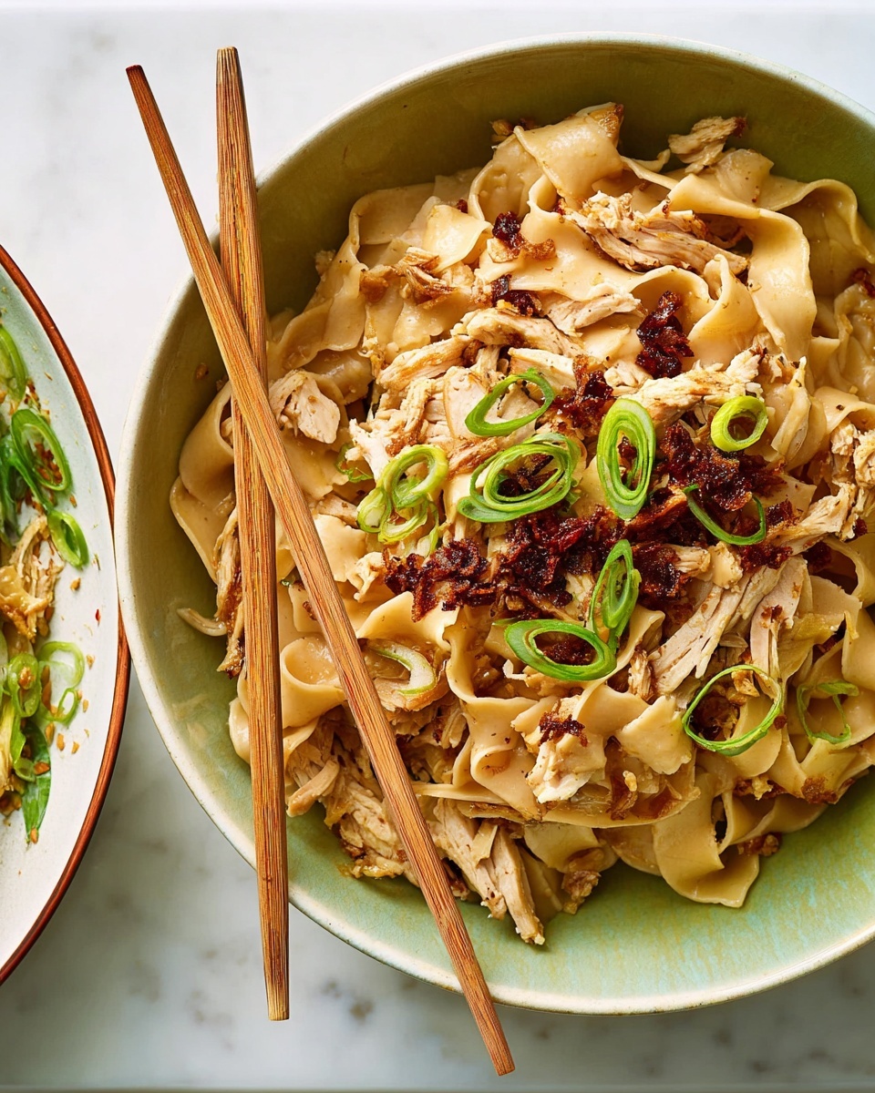 A close-up of a beige bowl filled with wide, flat noodles mixed with shredded chicken pieces and crispy brown bits scattered throughout. Thinly sliced green onions are sprinkled on top, adding a fresh green color contrast. The beige noodles look smooth and slightly glossy. Resting on the bowl are light wooden chopsticks. The bowl is placed on a white marbled surface, and part of another white bowl containing a colorful salad with greens, pomegranate seeds, and slices of beetroot is visible at the right edge. Photo taken with an iphone --ar 4:5 --v 7