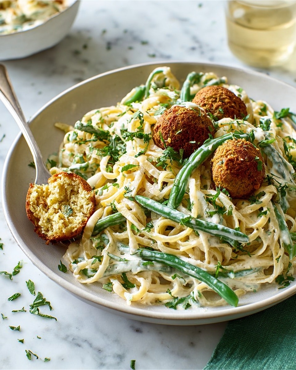 The image shows a white bowl filled with creamy fettuccine pasta mixed with green beans, both coated in a white sauce with specks of green herbs. On top and around the pasta, there are several brown, crispy-looking round falafel pieces, one of which is cut open, showing a beige inside. The pasta strands are tangled and slightly glossy from the sauce. Some chopped green herbs are sprinkled over everything, giving it a fresh look. The bowl is placed on a white marbled surface with a blurred glass of light-colored liquid and a small white bowl in the background, and a green cloth is partially visible on the side. A silver fork rests in the bowl. photo taken with an iphone --ar 4:5 --v 7