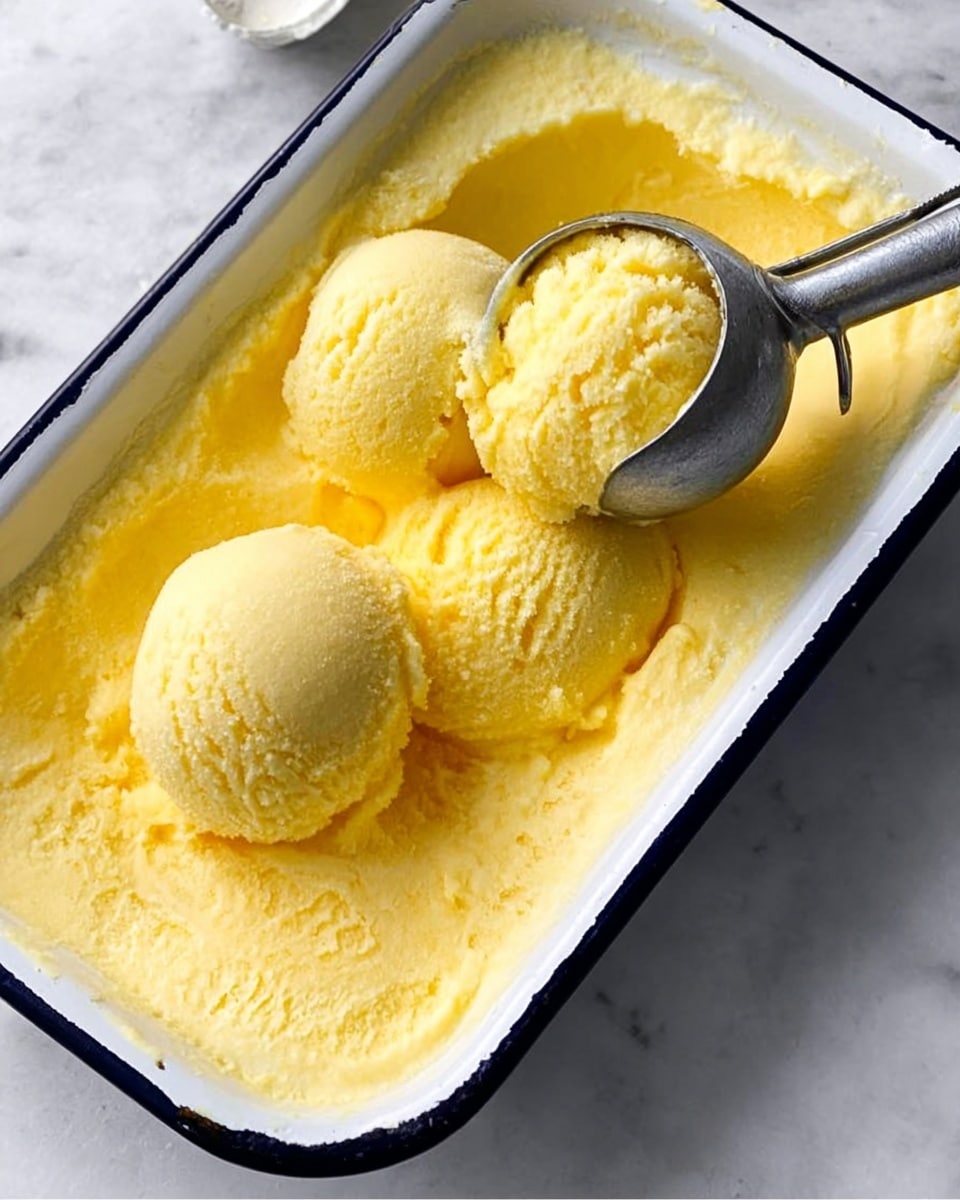 A white rectangular dish with a black rim holds a bright yellow, smooth, creamy frozen dessert with a soft texture. Three round scoops of the dessert sit together in the top half of the container, with one scoop partly lifted by a metal ice cream scooper. The inside surface of the dish shows smoothed and scraped areas of the dessert, revealing its thick consistency. The dish rests on a white marbled surface. Photo taken with an iphone --ar 4:5 --v 7