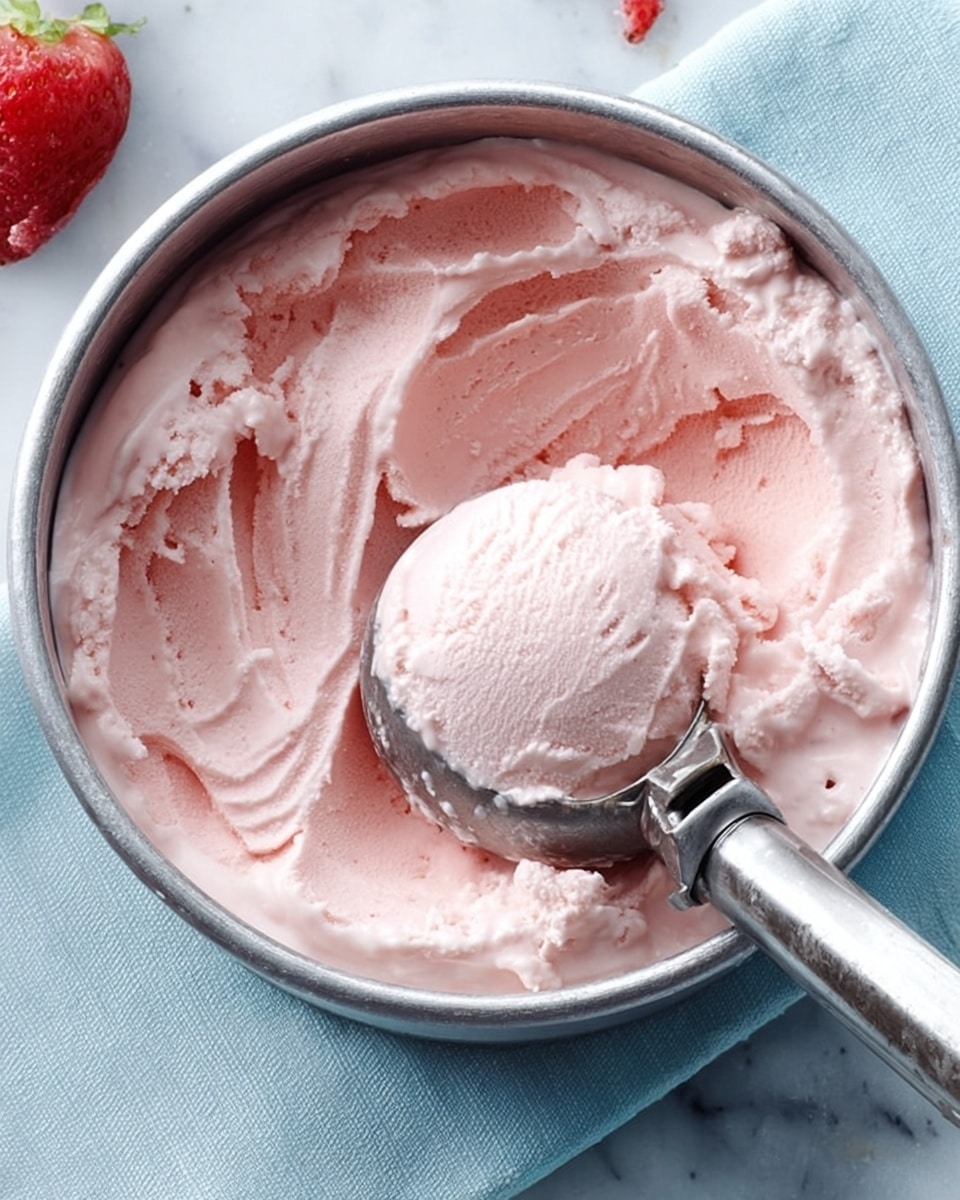 A close-up view of a metal bowl filled with smooth, light pink ice cream. The ice cream shows a creamy texture with some swirled and scooped areas, indicating softness. A shiny silver ice cream scoop with a ribbed handle rests inside the bowl, partially pressed into the ice cream. The bowl sits on a soft blue cloth, placed on a white marbled surface background. photo taken with an iphone --ar 4:5 --v 7