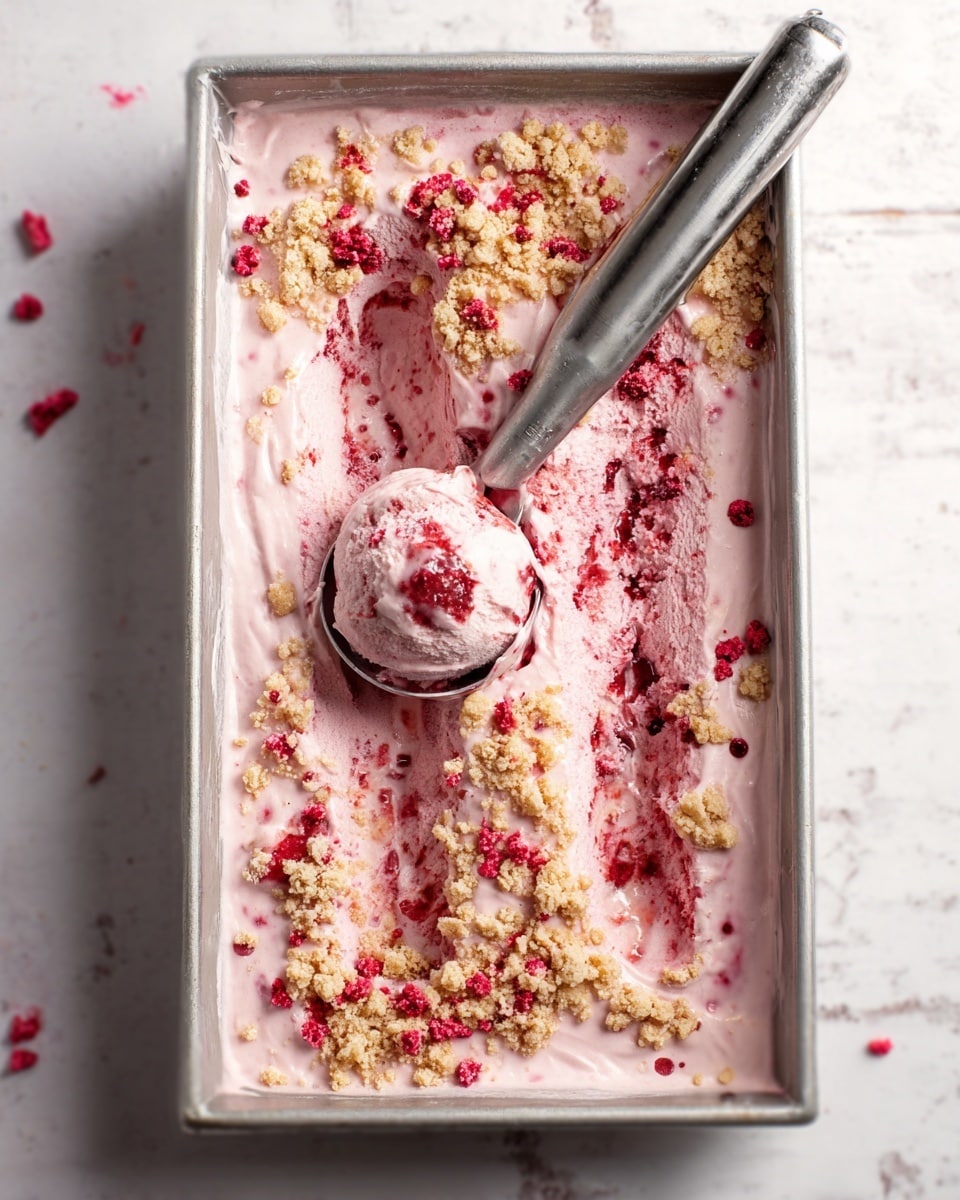 A rectangular metal container holds a pink ice cream mixed with crushed red berries spread unevenly on top, creating a rough texture with patches of creamy pink surface visible beneath. Scattered small crumbles of light beige cookie or crumble topping cover the surface, mainly along the edges. A large metal ice cream scoop with a smooth handle is resting inside, scooping a rounded portion of the ice cream, showing a soft and creamy texture with some berry bits inside. The container is placed on a white marbled surface. photo taken with an iphone --ar 4:5 --v 7
