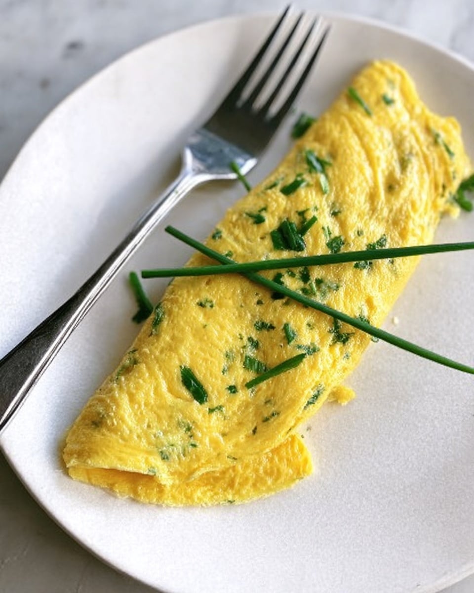 A yellow omelette with green herb pieces folded neatly on a white plate, its surface smooth and slightly shiny, with a few fresh green chives laid on top in a crisscross pattern. A silver fork is placed to the side of the omelette, resting on the plate. The background and surface are white with a marbled texture. Photo taken with an iphone --ar 4:5 --v 7