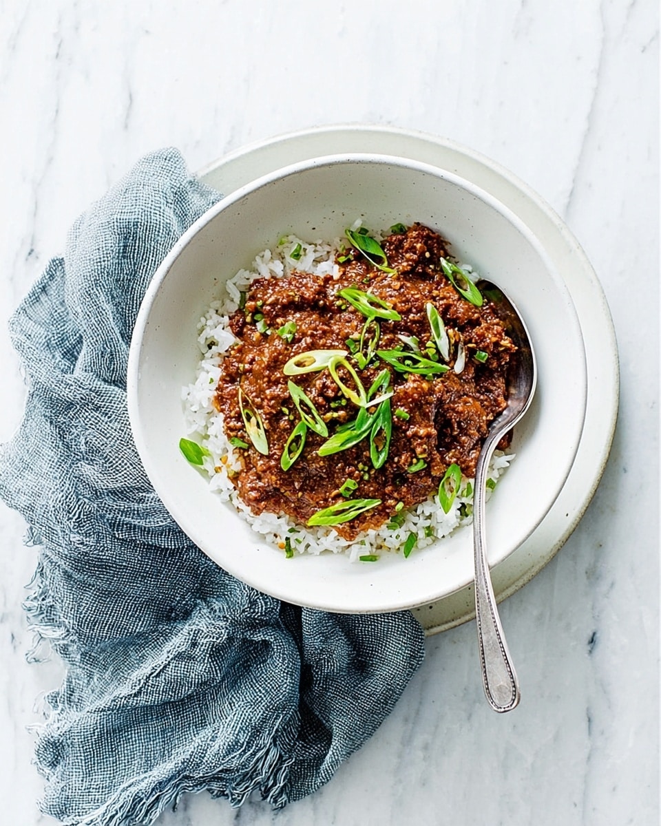 A white bowl filled with two main layers: a bottom layer of white rice with a soft, slightly fluffy texture, and a top layer of dark brown cooked ground meat with some red seasoning visible. Scattered green sliced spring onions sit on top of the meat, adding a fresh touch. A silver spoon rests inside the bowl on the left side. The bowl is placed on a white plate, with a light blue cloth napkin folded next to it on a white marbled surface. Photo taken with an iphone --ar 4:5 --v 7
