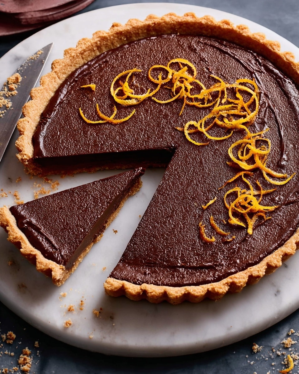 A round tart with a golden-brown crust holding a rich, dark brown chocolate filling appears on a white plate. The smooth chocolate surface is decorated with thin, curly orange zest strips scattered evenly on top. A single triangular slice has been cut out, revealing the dense chocolate inside, maintaining the same dark color as the top layer. The crust is thick and crimped along the edges, contrasting with the softer chocolate layer. A knife with chocolate remnants rests beside the tart on the plate, all set on a white marbled surface. photo taken with an iphone --ar 4:5 --v 7