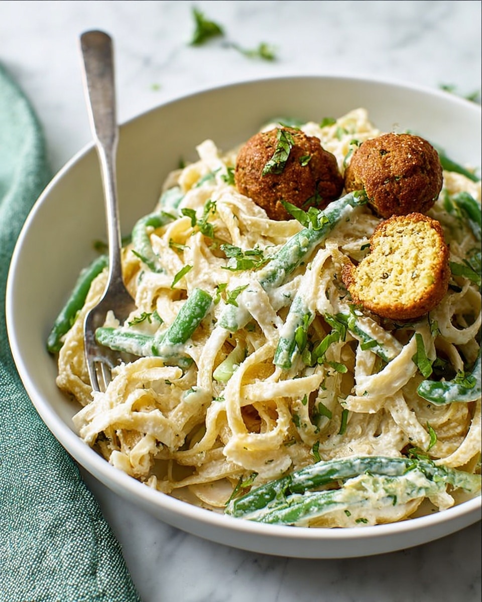 A white bowl filled with creamy fettuccine pasta mixed with green beans and sprinkled with chopped green herbs. On top and mixed in, there are several golden-brown falafel balls with a crispy texture, one of which is cut open inside the bowl showing its soft, crumbly center. The creamy white sauce coats the pasta and beans evenly, giving a rich look. A silver fork is resting on the left side of the bowl, placed on a white marbled surface with a folded green cloth nearby. photo taken with an iphone --ar 4:5 --v 7
