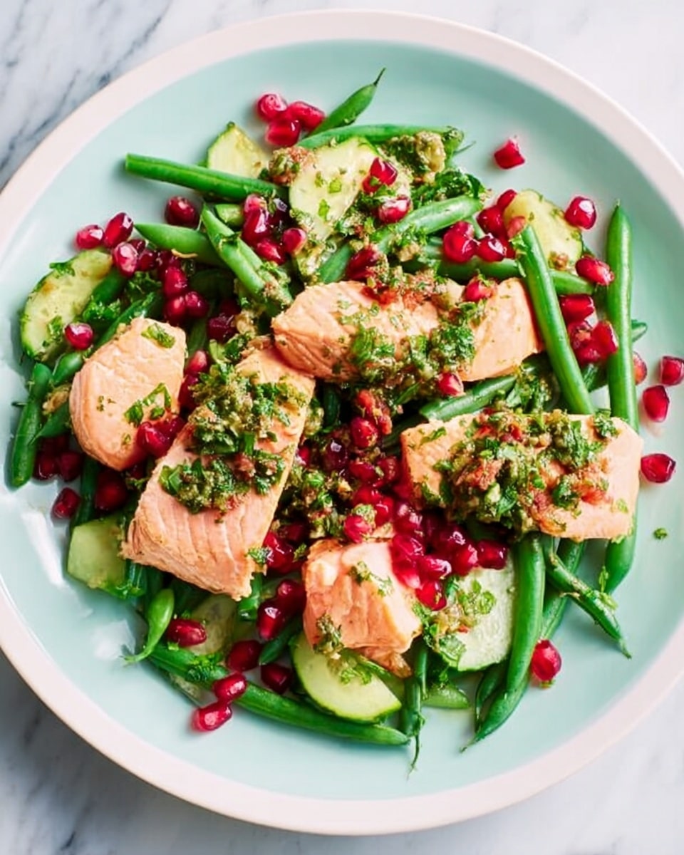 A white plate filled with a colorful dish featuring three pieces of cooked fish topped with a green herb sauce. Around the fish are bright green beans and slices of cucumber, with bright red pomegranate seeds scattered throughout. The dish also includes a grain-like base mixed with chopped herbs and small nuts. The plate is set on a white marbled surface with a blue background. photo taken with an iphone --ar 4:5 --v 7