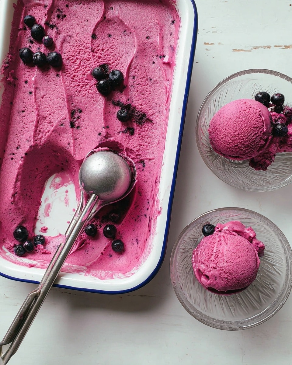 The image shows a white enamel tray with a blue rim filled with a bright purple creamy dessert, likely berry ice cream or sorbet, with a smooth texture and a few dark berries placed on top for decoration. Next to the tray, a shiny metal ice cream scoop with purple residue rests on a white marbled surface. Two clear glass dessert bowls hold one scoop each of the same purple ice cream, topped with small dark berries. The setting is bright, clean, and simple, focusing on the vibrant color contrast of the dessert against the white marbled background. Photo taken with an iphone --ar 4:5 --v 7