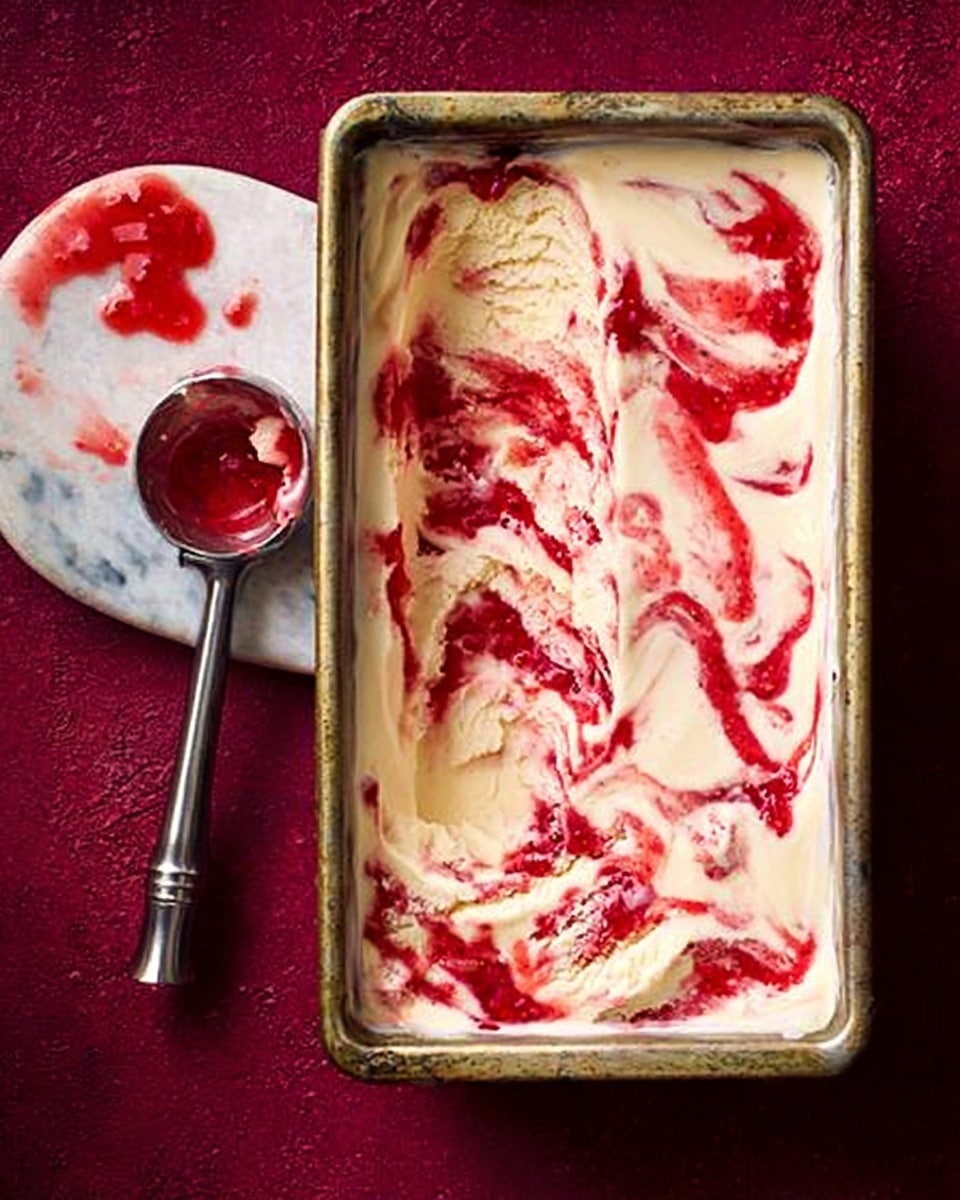 A rectangular metal pan filled with a creamy, pale yellow ice cream or dessert base, swirled richly with bright red strawberry or raspberry sauce, creating a marbled effect on top. The swirls are uneven and thick, spreading across the entire surface with some areas having more concentrated red color. Next to the pan, on the left, is a metal ice cream scoop resting on a white marbled surface, containing remnants of the dessert and red sauce. Small drops of sauce and cream are also scattered near the scoop. The image background is a deep reddish-purple color. Photo taken with an iphone --ar 4:5 --v 7