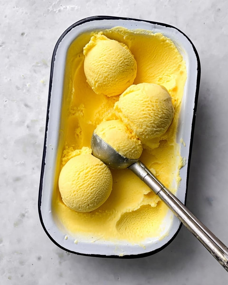 A white rectangular dish with a black rim filled with bright yellow ice cream. Three scoops of the yellow ice cream are placed on one side of the dish, with a metal ice cream scooper resting partially inside the ice cream next to the scoops. The texture of the ice cream is smooth but slightly soft, with some small rough patches where it has been scooped. The dish is on a white marbled surface. photo taken with an iphone --ar 4:5 --v 7