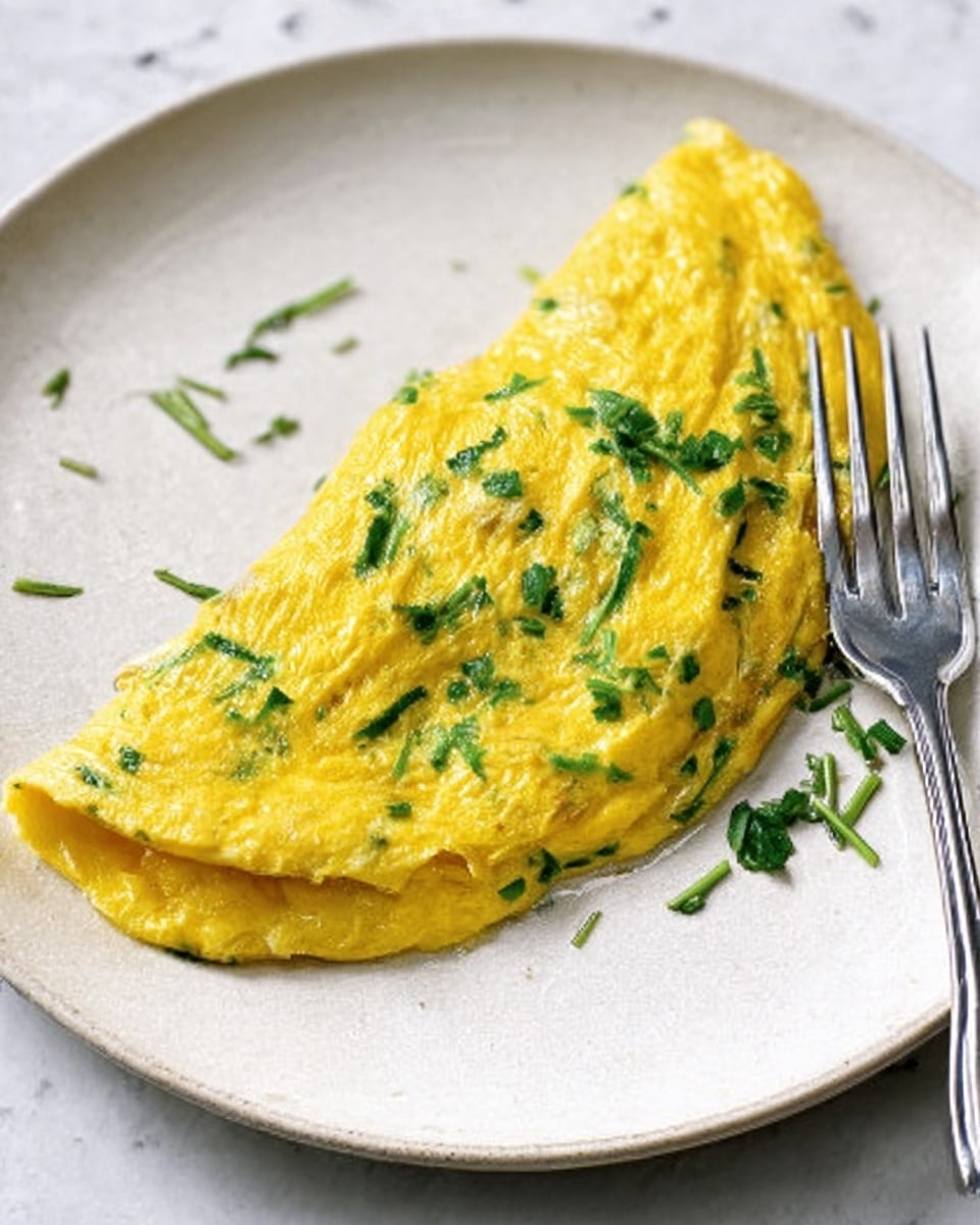 A folded omelet sits on a clean white plate over a white marbled surface, showing a smooth yellow outer layer with small green specks of herbs mixed in. The omelet is thick and slightly soft in texture, with two thin green chives placed crosswise on top for garnish. A silver fork lies partially under the omelet on the lower left side, its shiny surface reflecting light. The overall scene is simple, bright, and fresh. photo taken with an iphone --ar 4:5 --v 7