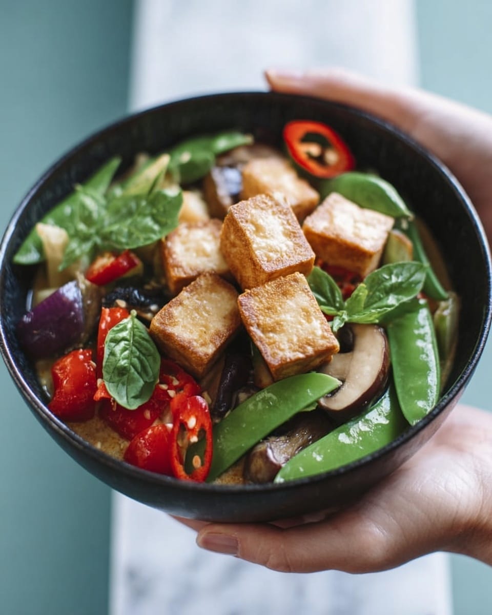 A white bowl filled with three main layers of colorful ingredients is being held by a woman's hand. The bottom layer shows a light green curry sauce with a smooth texture. On top are chunky pieces of browned tofu, green snap peas, red chili slices, light brown mushrooms, purple eggplant pieces, and red bell pepper chunks. Fresh green basil leaves are scattered on top, adding a fresh touch. The bowl is held above a white marbled surface. photo taken with an iphone --ar 4:5 --v 7