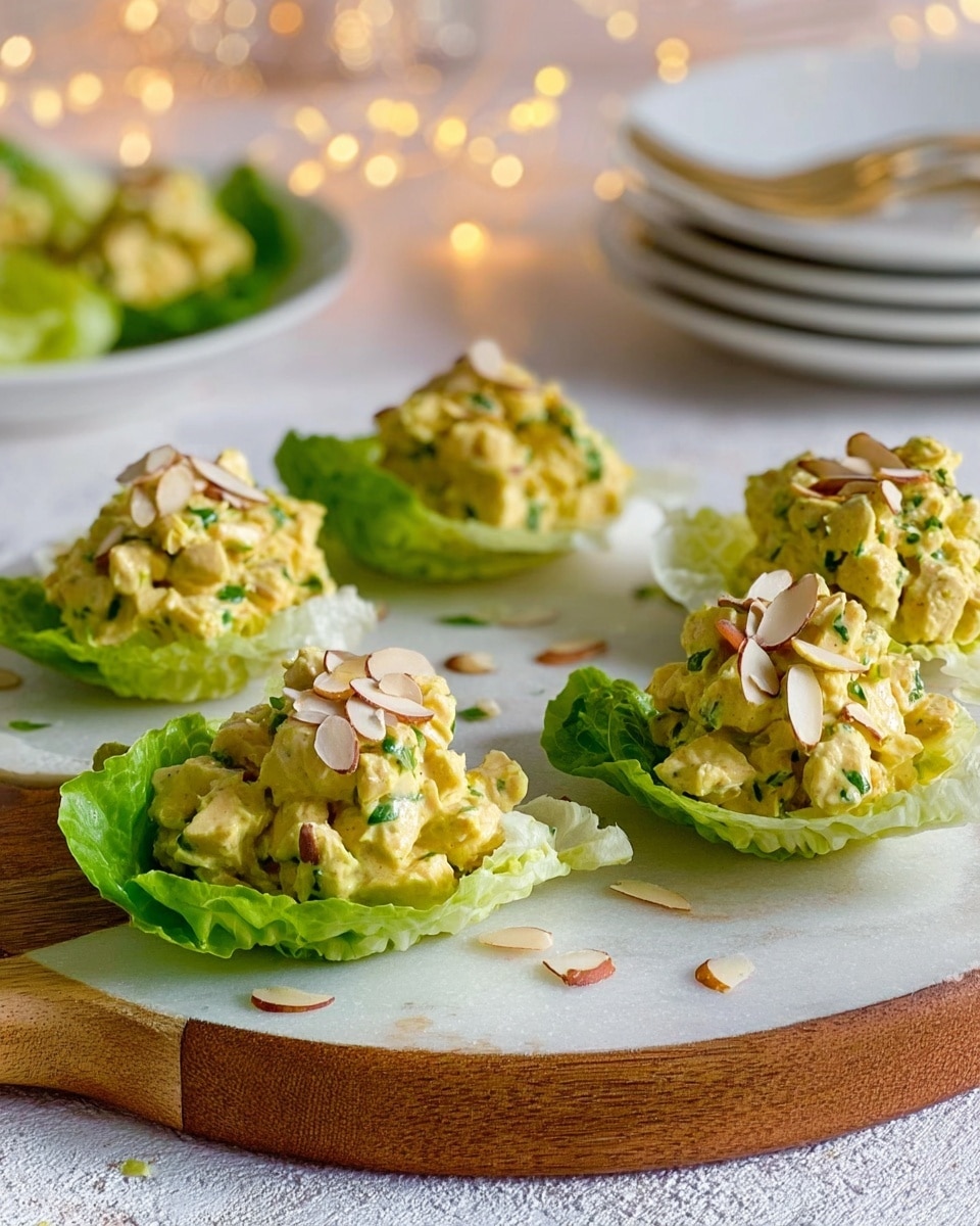 The image shows five lettuce cups arranged on a round white serving board placed on a white marbled surface. Each cup is made with a bright green lettuce leaf that holds a filling of yellow chicken salad mixed with small green herbs and topped with light brown almond slices. Some almond slices are also scattered on the board and the marbled surface around it. Soft white fairy lights and a stack of white plates with more lettuce cups in the background add a cozy feel to the scene. photo taken with an iphone --ar 4:5 --v 7