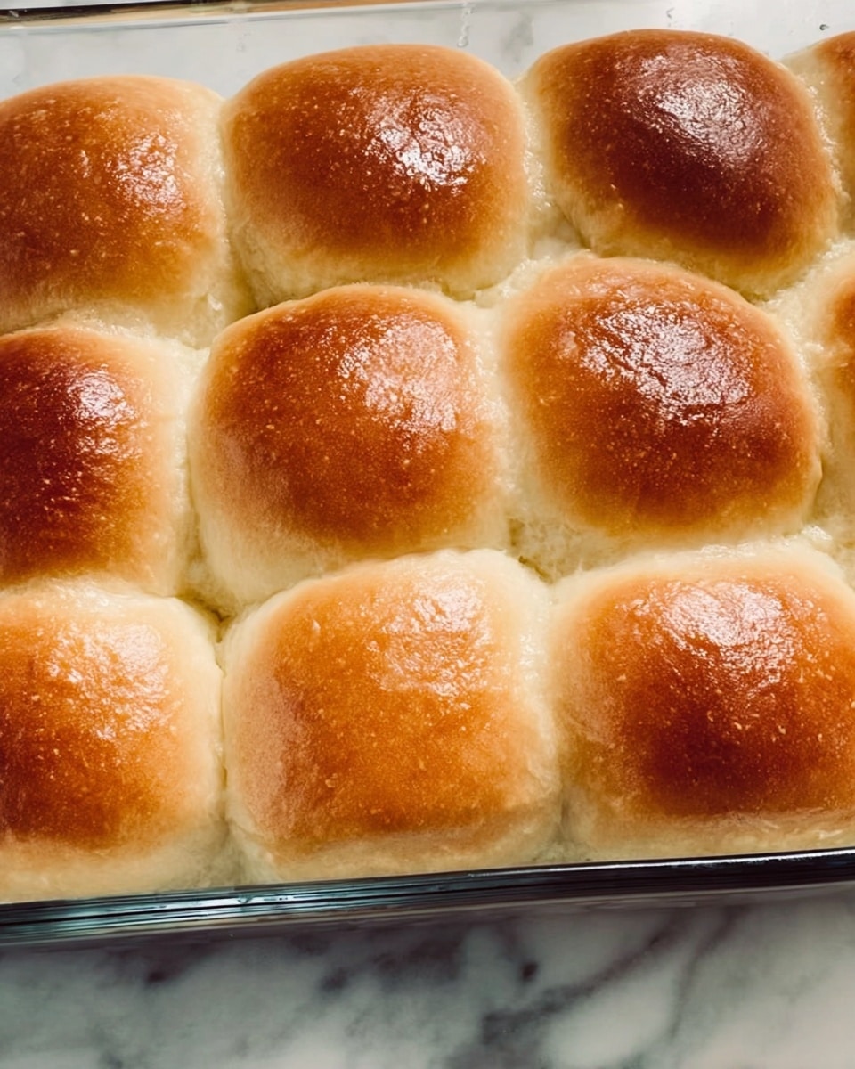 This image shows a close-up of a glass baking dish filled with nine golden-brown dinner rolls arranged in a 3 by 3 grid. Each roll has a smooth, shiny top with a slightly darker brown center, showing a well-baked texture. The rolls are touching each other, making a soft, puffy surface, and the edges of the dish show some slight browning. The baking dish rests on a white marbled surface. photo taken with an iphone --ar 4:5 --v 7