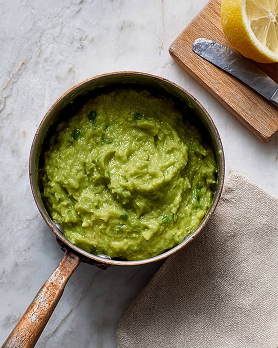 The image shows a small metal pot filled with a thick, chunky green sauce that looks like guacamole, placed on a white marbled surface. The sauce has a creamy texture with visible small bits of avocado and herbs giving it a fresh, uneven surface. Next to the pot is a wooden cutting board with a cut lemon, its inside bright yellow, and a metal knife lying beside it. A light beige cloth napkin is partially seen under the pot. The whole scene is lit with soft natural light, making the colors rich yet gentle. photo taken with an iphone --ar 4:5 --v 7