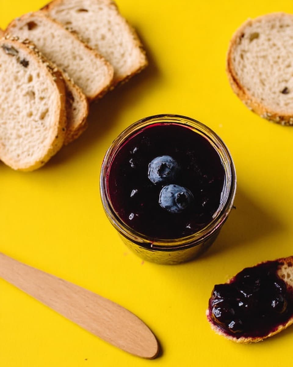 The image shows a jar of dark purple blueberry jam with two whole blueberries on top, placed on a white marbled surface. Above the jar, there are three pieces of light brown toasted bread stacked loosely. To the right, a small round slice of bread is spread with the same dark purple jam. A small wooden spreader lies diagonally next to the jam-covered bread slice. The background is a bright yellow color, making the jam and bread stand out clearly. photo taken with an iphone --ar 4:5 --v 7