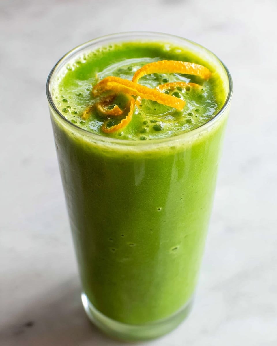 A tall clear glass filled with a thick green smoothie, showing a slightly frothy texture on top. The surface has swirls of light green and darker green with a few bubbles visible. Three thin orange peels are curled and placed gently on the smoothie's top surface. The glass is set on a white marbled texture. photo taken with an iphone --ar 4:5 --v 7