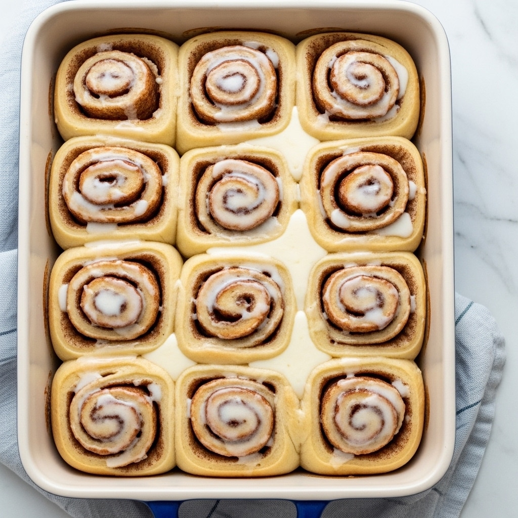 A white baking dish filled with 14 golden brown cinnamon rolls arranged in three rows, each roll showing a clear spiral pattern with a light glaze spread unevenly on top. The dish is partly covered with a light blue and white cloth on a white marbled surface. The glaze adds a slightly shiny, creamy texture contrasting with the soft, fluffy rolls below. Photo taken with an iphone --ar 4:5 --v 7