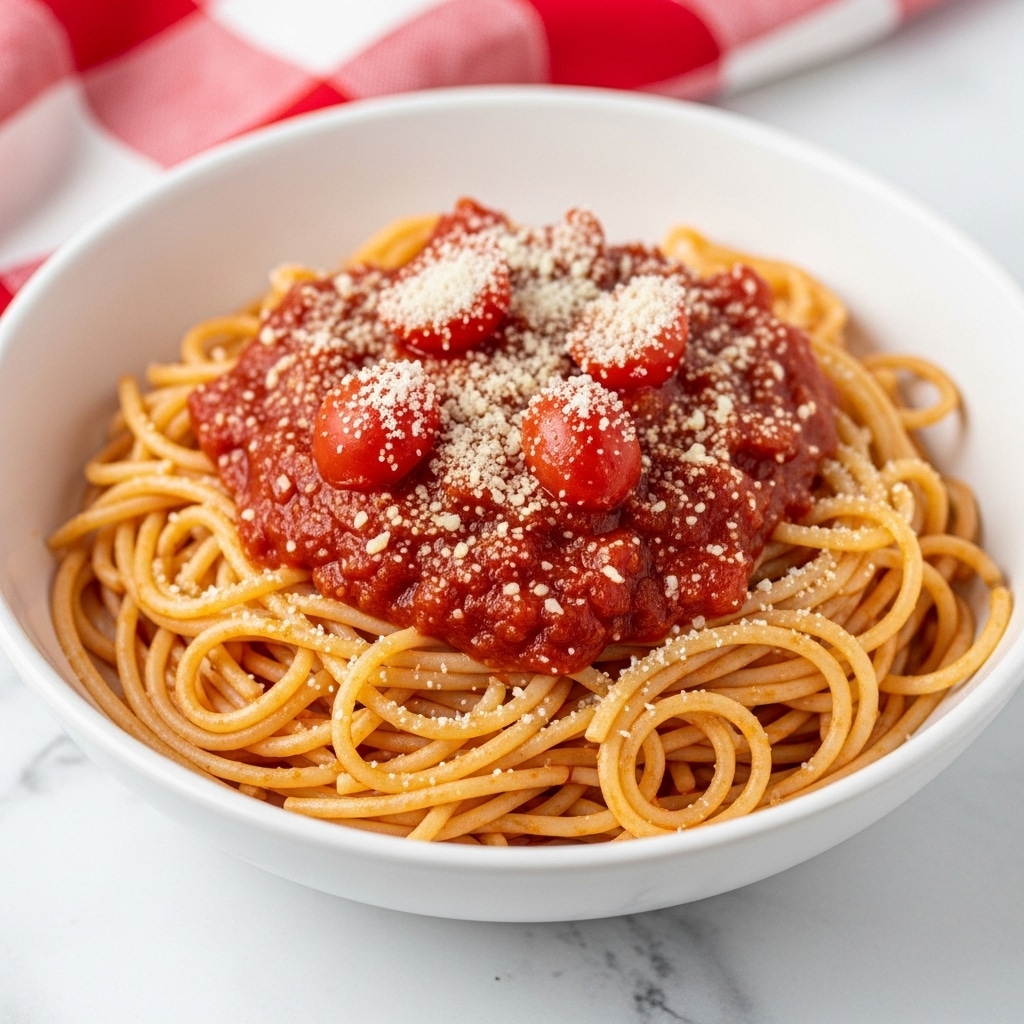 A white bowl holds a neat swirl of spaghetti coated in a bright red tomato sauce with small chunks of tomato on top. The pasta is topped with a light dusting of grated cheese, adding a soft white texture over the rich, glossy red sauce. The bowl sits on a white marbled surface with a red and white checkered cloth partially visible around it. photo taken with an iphone --ar 4:5 --v 7