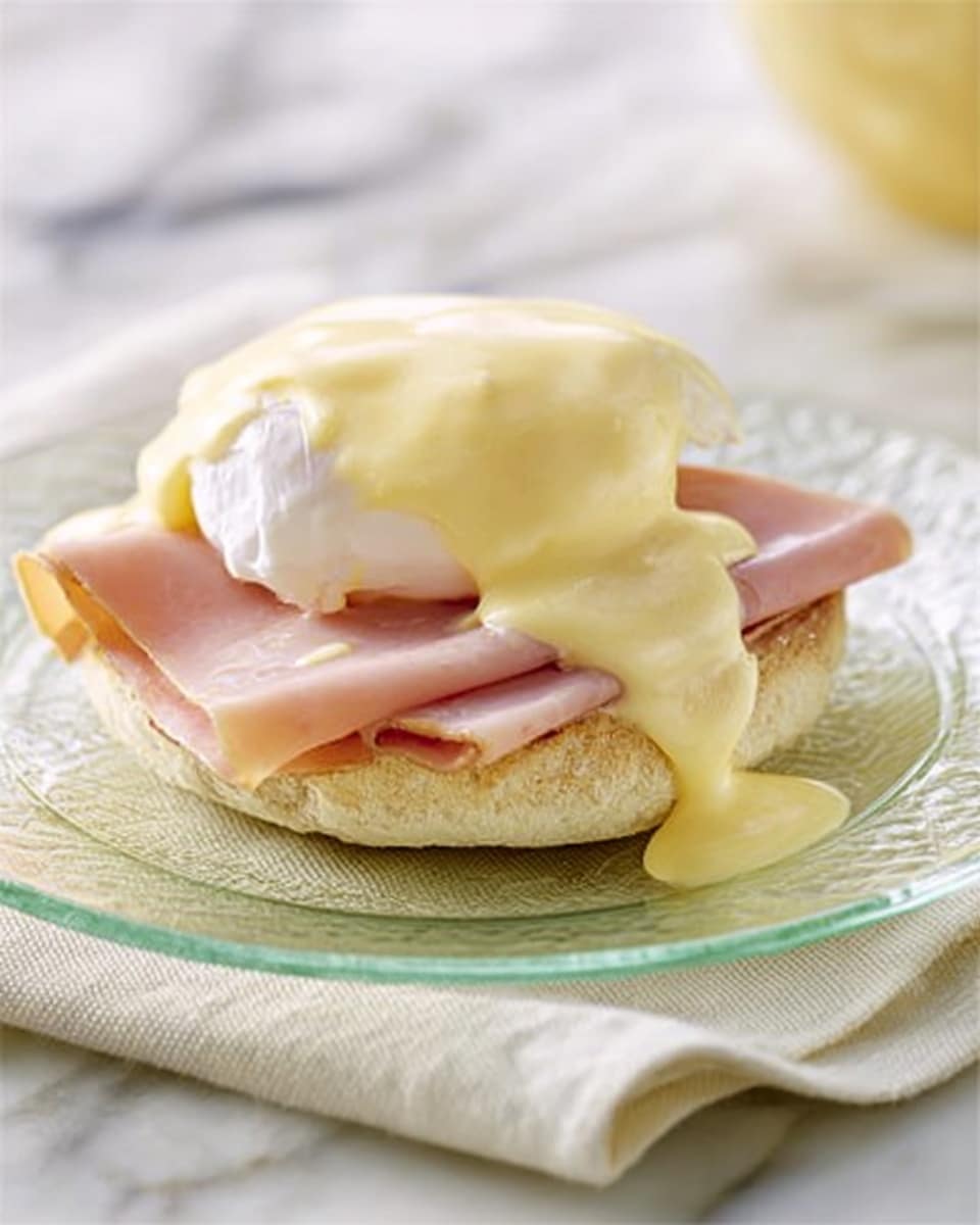 A soft English muffin sits at the base, its golden-brown toasted surface visible around the edges. On top of the muffin is a thin layer of folded pink ham, slightly ruffled. Above the ham rests a smooth, white poached egg with a shiny surface. The whole stack is covered by a thick, creamy pale yellow hollandaise sauce that drips slightly over the sides of the muffin. The dish is served on a clear glass plate placed on a white cloth, which lies on a white marbled surface. Photo taken with an iphone --ar 4:5 --v 7