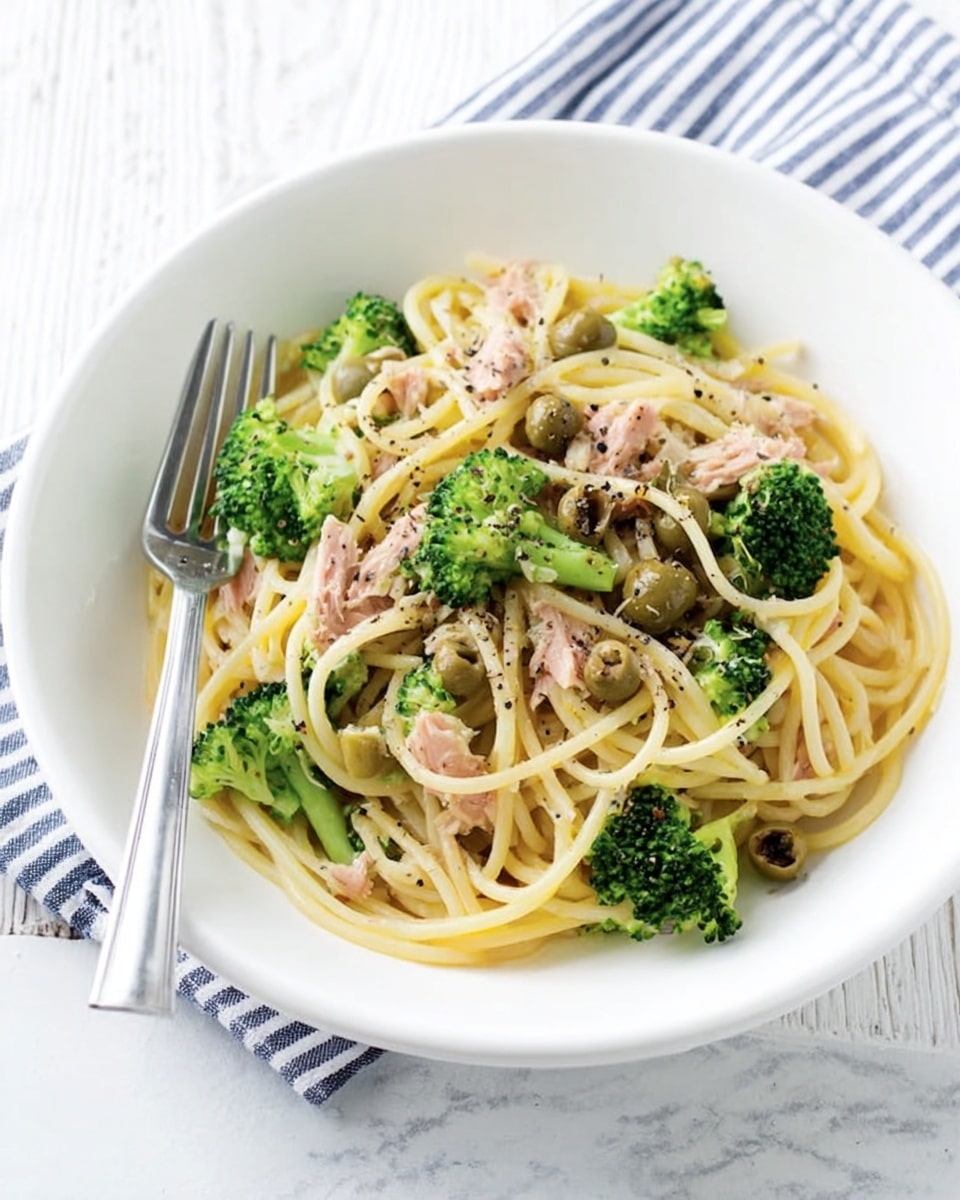 A white bowl filled with spaghetti pasta mixed with green broccoli florets, sliced olives, and small pieces of tuna scattered evenly throughout. The spaghetti strands are light yellow and slightly shiny, showing a soft texture. The broccoli is bright green and fresh, adding a pop of color. The olives are light brown with a smooth surface, blending with the tuna chunks, which are a muted pink color with soft texture. The dish is lightly sprinkled with black pepper, adding tiny black dots mainly on the pasta. A silver fork rests inside the bowl on the left side. The bowl sits on a white marbled surface with a faint striped cloth in blue and white in the background. Photo taken with an iphone --ar 4:5 --v 7