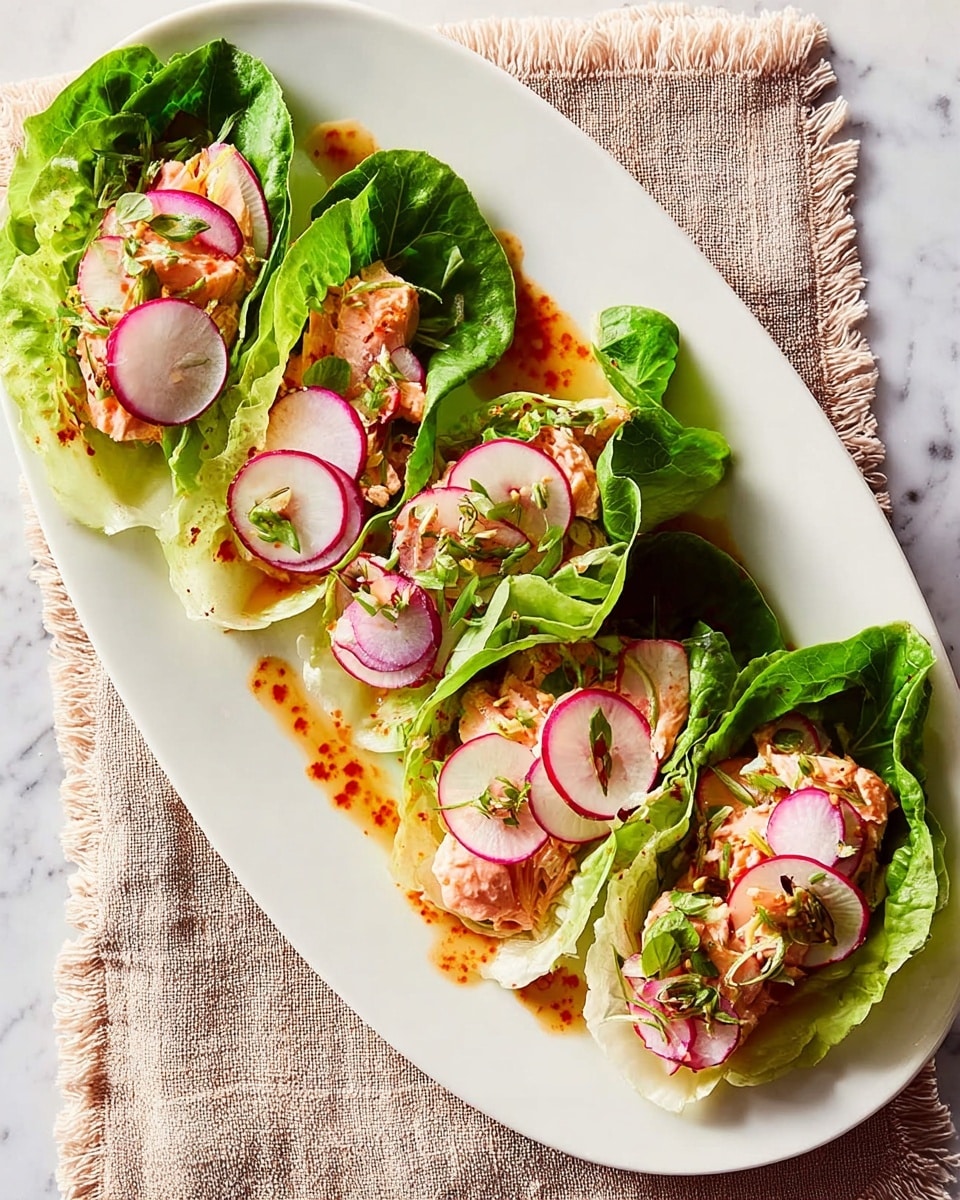 The image shows five small lettuce wraps arranged on a large white oval plate. Each wrap has a base layer of bright green lettuce leaves, topped with light pink pieces of cooked salmon. On top of the salmon are thin, round slices of white and purple radishes, and light green slices of other vegetables. The wraps have a drizzle of red sauce over them and are garnished with small green herb pieces. The plate is placed on a light brown textured fabric, all set on a white marbled surface. photo taken with an iphone --ar 4:5 --v 7