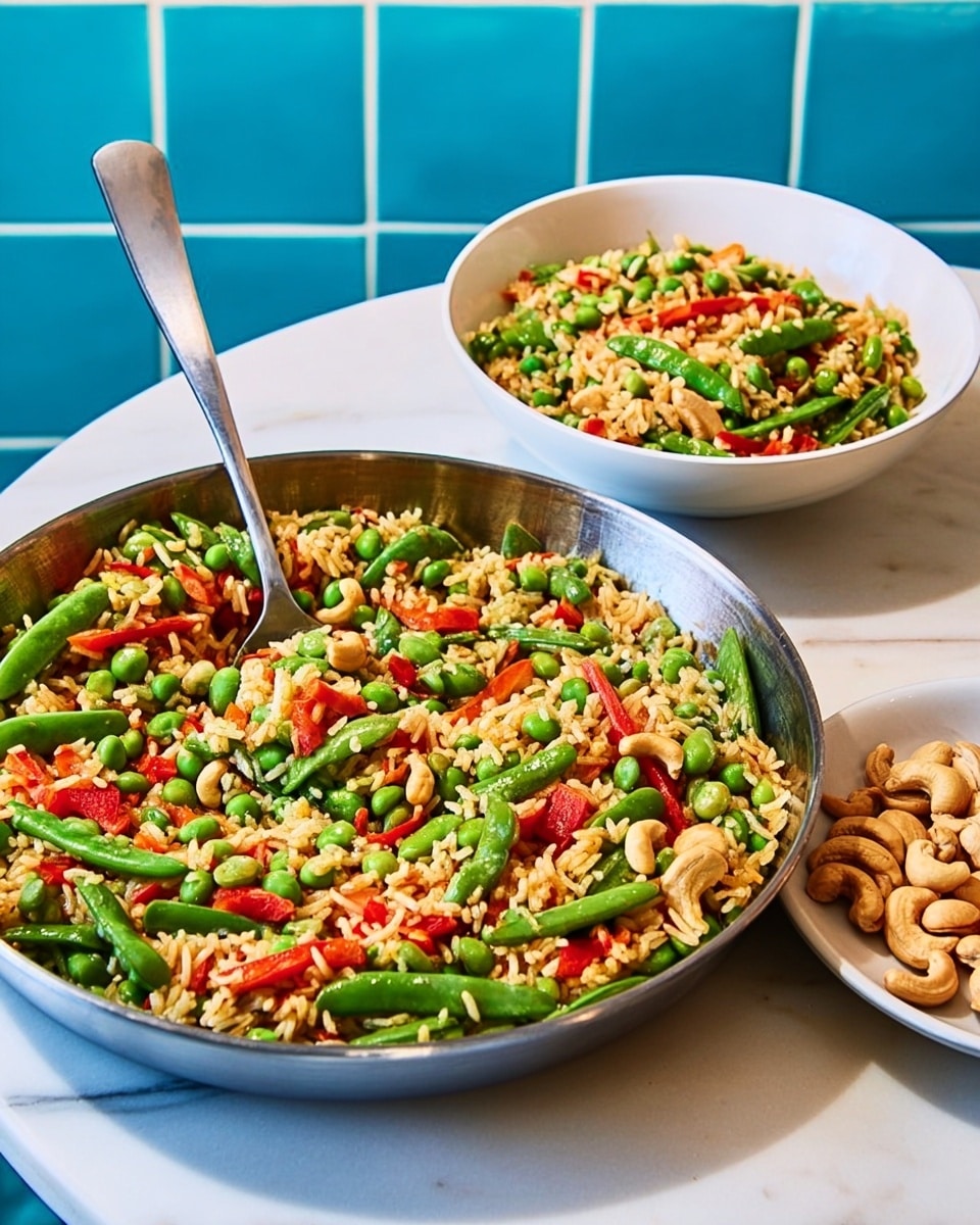 A large pan filled with a colorful mixed vegetable rice dish sits on a white marble surface, with the pan itself having a rustic, worn metal look and a long handle. The dish contains visible layers of white rice mixed with green beans, bright green edamame, and small pieces of red bell pepper scattered throughout. A metal spoon is resting inside the pan, partially scooping some of the food. On the side, there is a white plate with a similar serving of the same dish, and next to it a small blue square dish holds light-colored cashew nuts. The background features blue tiled walls, adding contrast to the white marble surface where all items are placed. Photo taken with an iphone --ar 4:5 --v 7