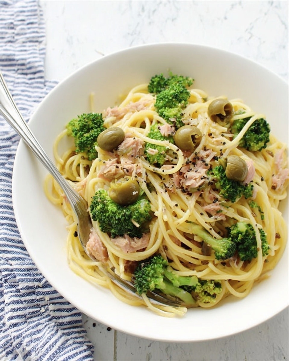 A white bowl holds a dish of spaghetti pasta mixed with green broccoli florets, light brown olives, and small pieces of light pink tuna. The pasta looks soft and is tossed with a light sauce. Black pepper is sprinkled over the top, adding a speckled dark touch. A silver fork rests on the left side of the bowl, slightly inside the pasta. The bowl is set on a white marbled surface with a white and blue striped cloth in the background. Photo taken with an iphone --ar 4:5 --v 7