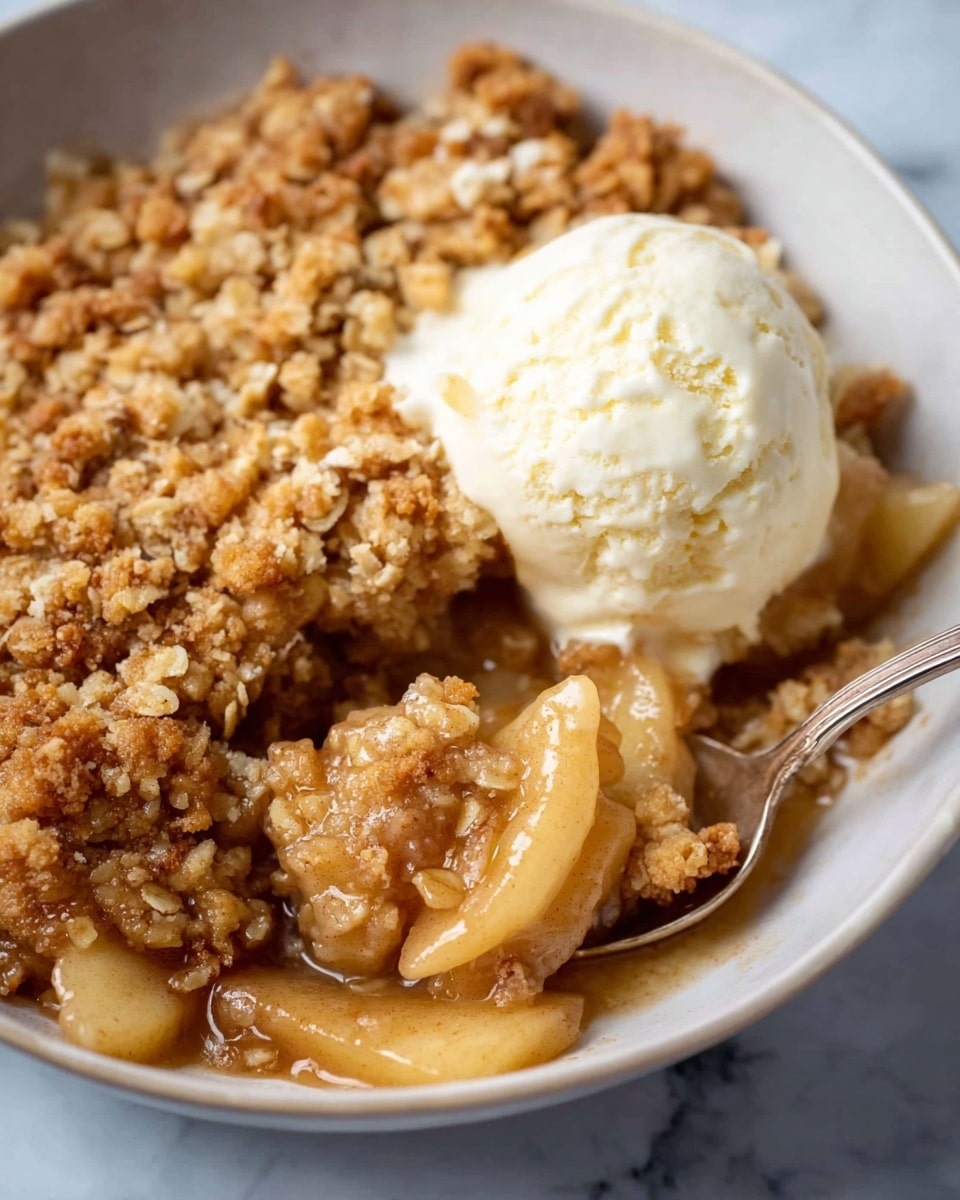 A close-up view of a white bowl filled with warm apple crumble dessert. The bottom layer shows soft, cooked apple pieces in syrup with a light brown color. On top, there is a thick, golden-brown crumbly oat topping, crispy and textured. A scoop of creamy vanilla ice cream with a smooth, pale off-white surface rests on one side of the crumble. A spoon is partially scooping the dessert, showing the layers clearly. The bowl is set on a white marbled surface. photo taken with an iphone --ar 4:5 --v 7