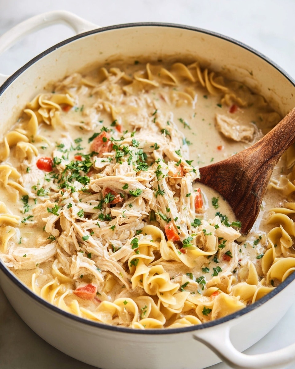 A close-up view of a black pot filled with creamy pasta. The dish has three main layers: the bottom layer is curly egg noodles in a light yellow color, followed by shredded pieces of cooked chicken mixed evenly, and scattered small diced red bell peppers throughout. The top layer is a thick, creamy white sauce surrounding the noodles and chicken, with green chopped herbs sprinkled over all. A wooden spoon partially dipped in the sauce rests on the right side of the pot. The pot is placed on a white marbled surface with some blurred greenery in the background. Photo taken with an iphone --ar 4:5 --v 7
