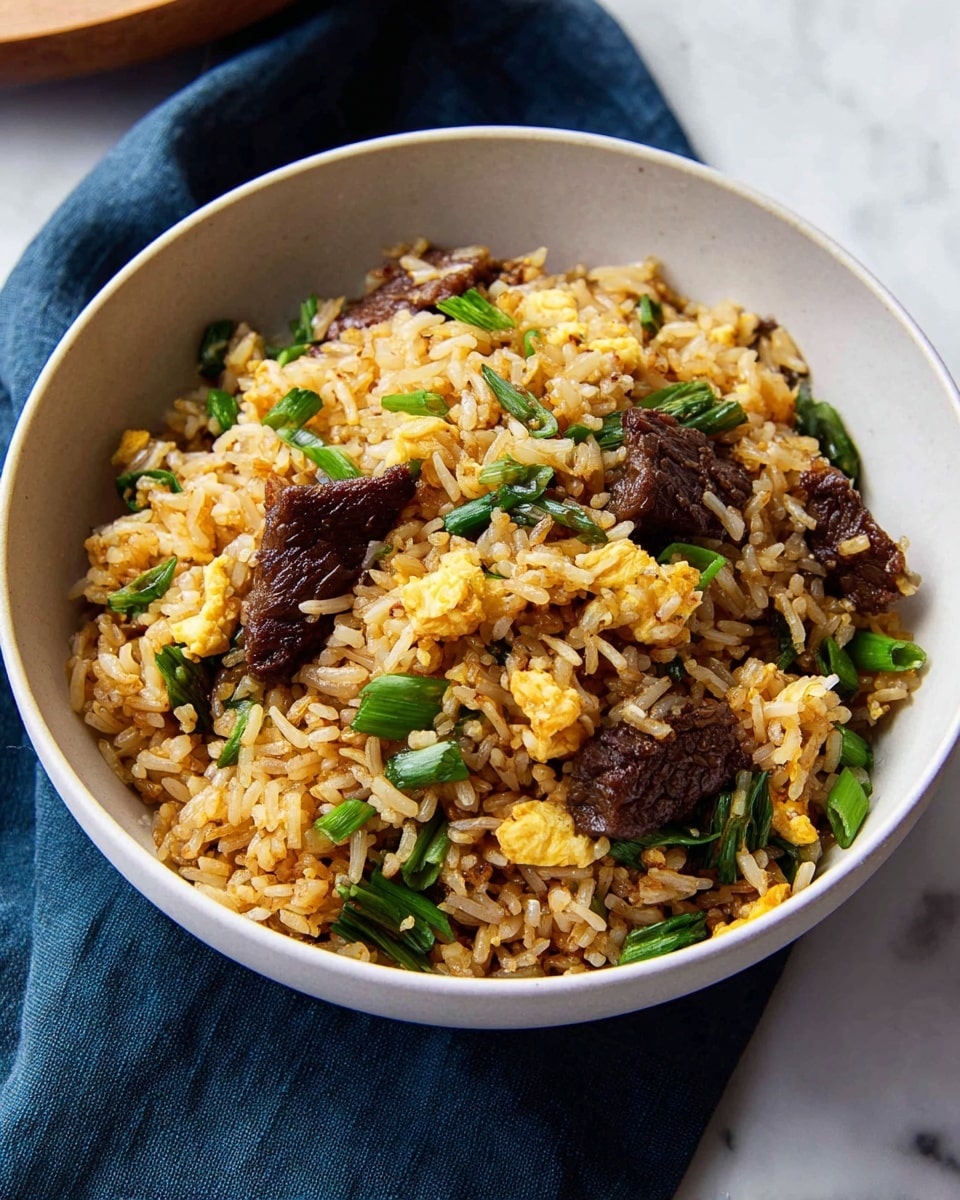 In a white bowl, there is one layer of fried rice with small, separate grains colored light brown from seasoning, mixed evenly with medium-sized, dark brown pieces of cooked beef, bright green chopped vegetables scattered throughout, and small bits of scrambled yellow egg. The bowl sits on a white marbled surface with a deep blue cloth partially underneath, giving contrast to the warm colors of the dish. In the background, the edge of another white bowl with the same fried rice is partially visible, slightly blurred. Photo taken with an iphone --ar 4:5 --v 7