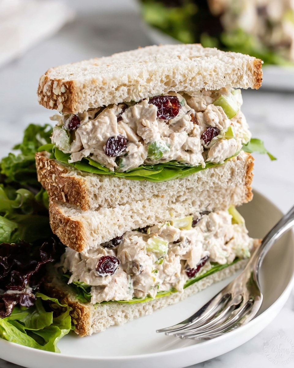 A sandwich cut in half and stacked with three layers: the top and bottom layers are thick slices of light brown bread with a soft texture, the middle layer contains green leafy lettuce with a fresh, ruffled texture, and inside is a creamy chicken salad mixed with diced celery and dried cranberries, giving a mix of white, green, and dark red colors. The sandwich halves rest on a white plate with mixed greens on the side, all placed on a white marbled surface. photo taken with an iphone --ar 4:5 --v 7