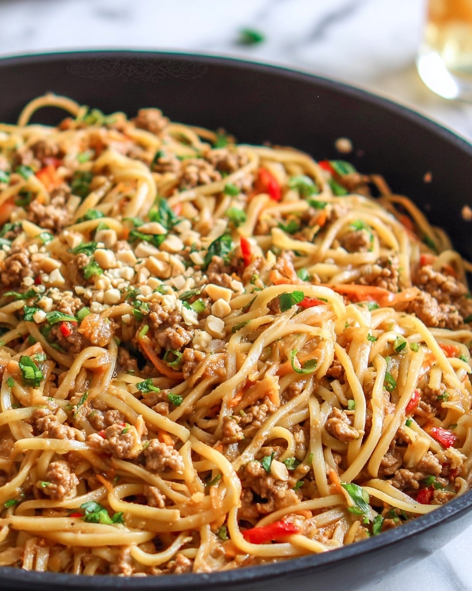 A close-up view of a black pan filled with cooked noodles mixed with small bits of ground meat and vegetables. The noodles are light brown and slightly glossy, twisted and tangled throughout the pan. Scattered on top are chopped green herbs, small pieces of red bell pepper, and sliced green onions, adding bright green and red colors. The dish looks saucy but not wet, with a rich and hearty texture. The background shows a soft white marbled texture with a hint of a blurred cloth in the distance. photo taken with an iphone --ar 4:5 --v 7