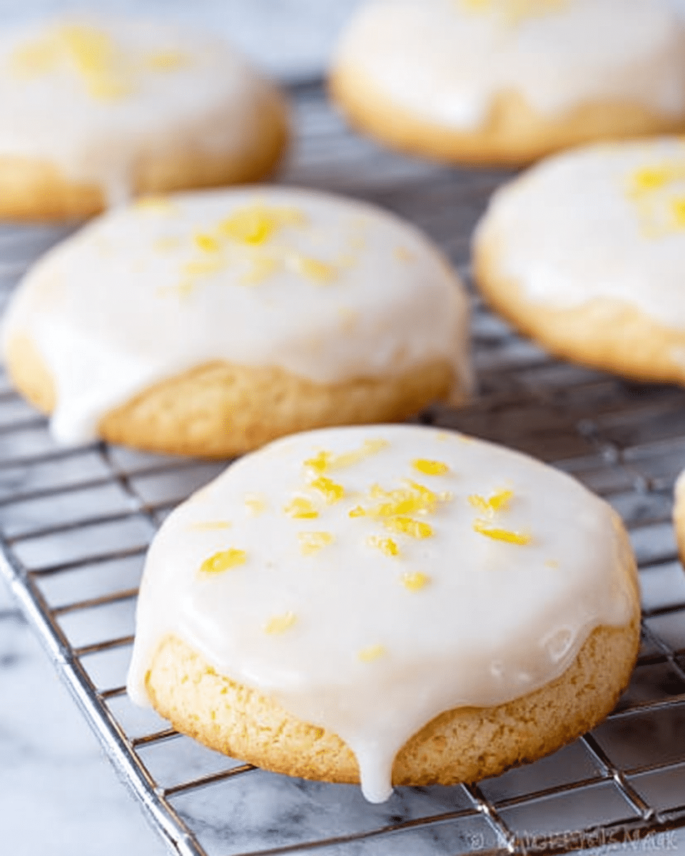 The image shows several round sugar cookies on a metal cooling rack, each cookie covered with a smooth, white glaze that drips slightly over the edges. The cookies have a light golden-brown color beneath the glaze, and there are tiny yellow crumbs or zest sprinkled on top of the glaze, adding a pop of color. The background is a white marbled surface, softly blurred to keep the focus on the cookies. Photo taken with an iphone --ar 4:5 --v 7