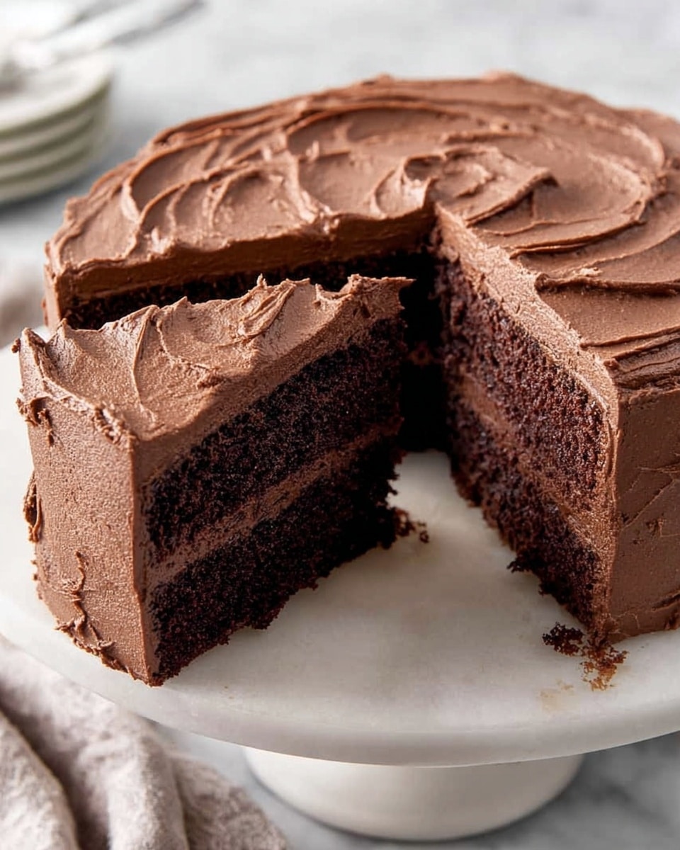 A round chocolate cake with two thick layers topped and covered in smooth, swirled chocolate frosting sits on a white cake stand. One slice is cut out and slightly lifted, showing the dark, moist chocolate cake layers separated by a thinner layer of the same chocolate frosting inside. The surface under the cake stand is a white marbled texture. Photo taken with an iphone --ar 4:5 --v 7