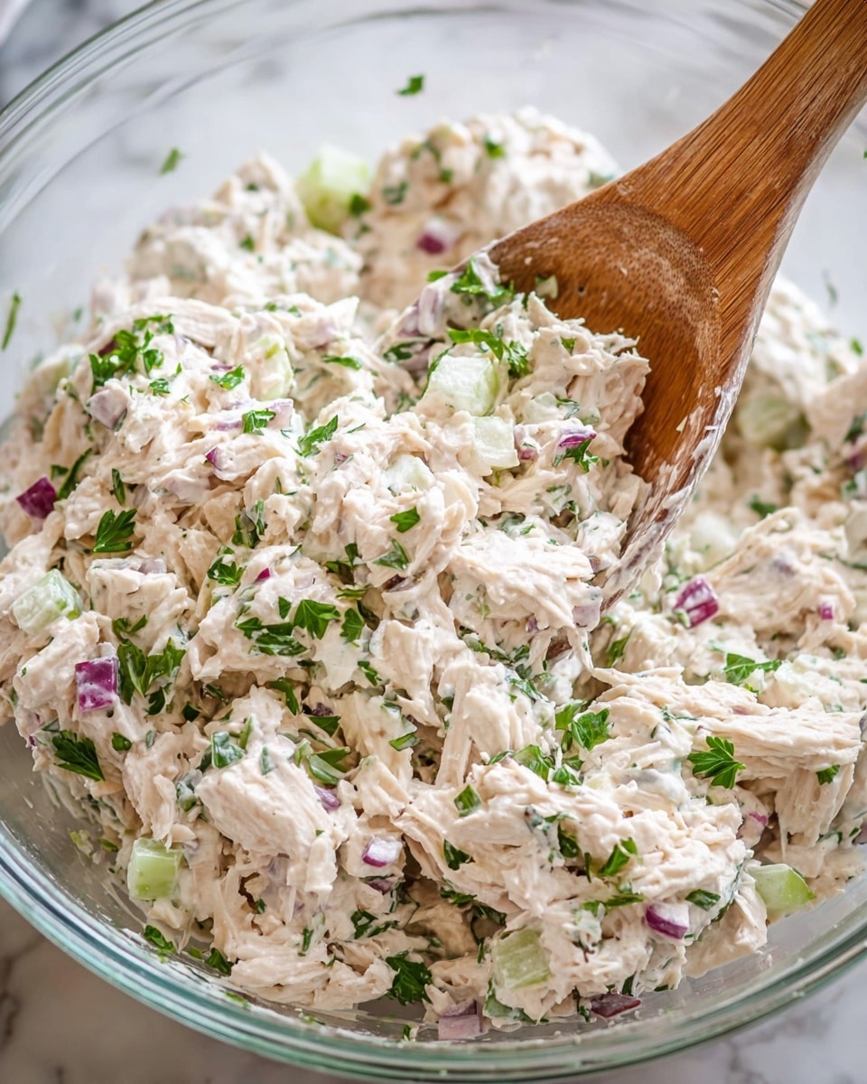 The image shows a clear glass bowl filled with a creamy chicken salad mix. The salad has several layers of shredded white chicken, diced light green celery, small pieces of purple-red onion, all lightly mixed with thick white dressing. There are small green parsley bits scattered throughout. A wooden fork is gently lifting some salad on the right side with dressing sticking to the tines. The bowl sits on a white marbled texture surface. photo taken with an iphone --ar 4:5 --v 7