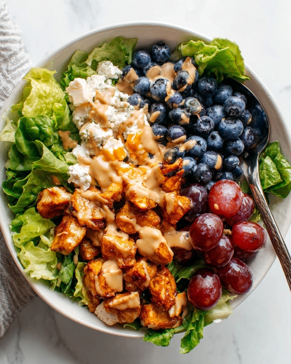 A white bowl filled with a colorful salad is set on a white marbled surface. The bowl contains four main layers: fresh green leafy lettuce on one side, a cluster of red grapes to the left, a portion of dark blueberries with a spoon on the upper right, and a creamy white cheese mixed with some orange bits on the bottom right. In the center, there are pieces of grilled chicken topped with a drizzle of light brown sauce, along with a sprinkling of chopped nuts. The textures range from smooth cheese, juicy fruit, tender chicken, and crunchy nuts. Photo taken with an iphone --ar 4:5 --v 7