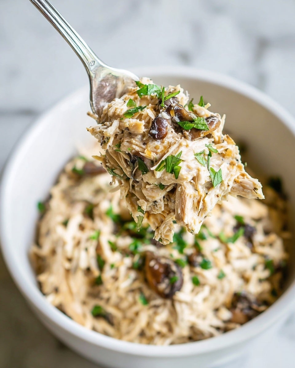 A close-up view of a creamy shredded chicken dish held on a metal spoon above a white bowl. The chicken is mixed with small chunks of caramelized onions and dark brown mushrooms, all coated in a creamy light beige sauce with visible black pepper specks. Bright green chopped herbs are scattered on top, adding a fresh color contrast. The white bowl sits on a white marbled surface. Photo taken with an iphone --ar 4:5 --v 7