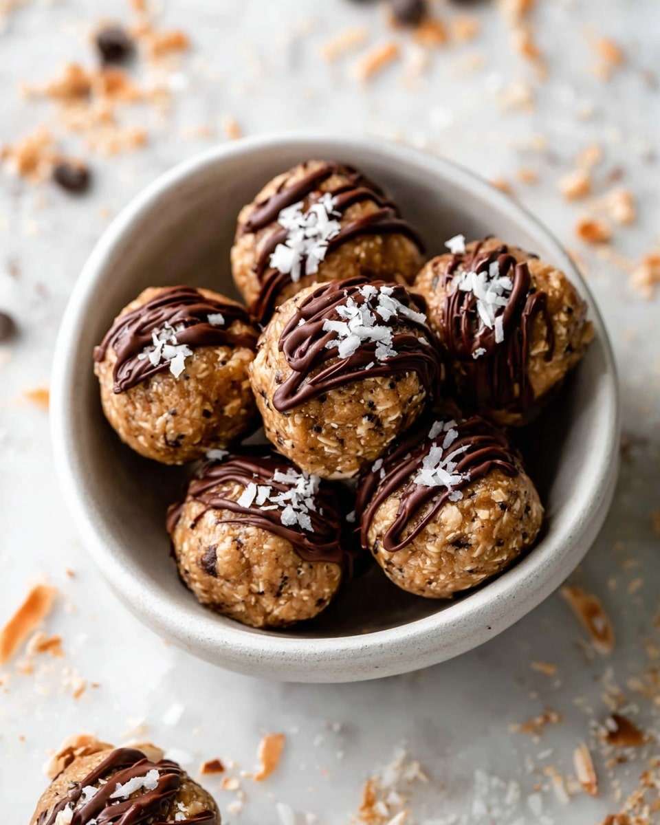 A white bowl filled with round energy bites that have a light brown color with small dark chia seeds visible. Each bite has a layer of dark chocolate partially covering its bottom half, with additional chocolate drizzled over the top, creating thin wavy lines. On top of the bites, there are small pieces of shredded coconut adding texture and light color contrast. The bowl is placed on a white marbled surface with some scattered bits of chocolate and coconut around it. Photo taken with an iphone --ar 4:5 --v 7