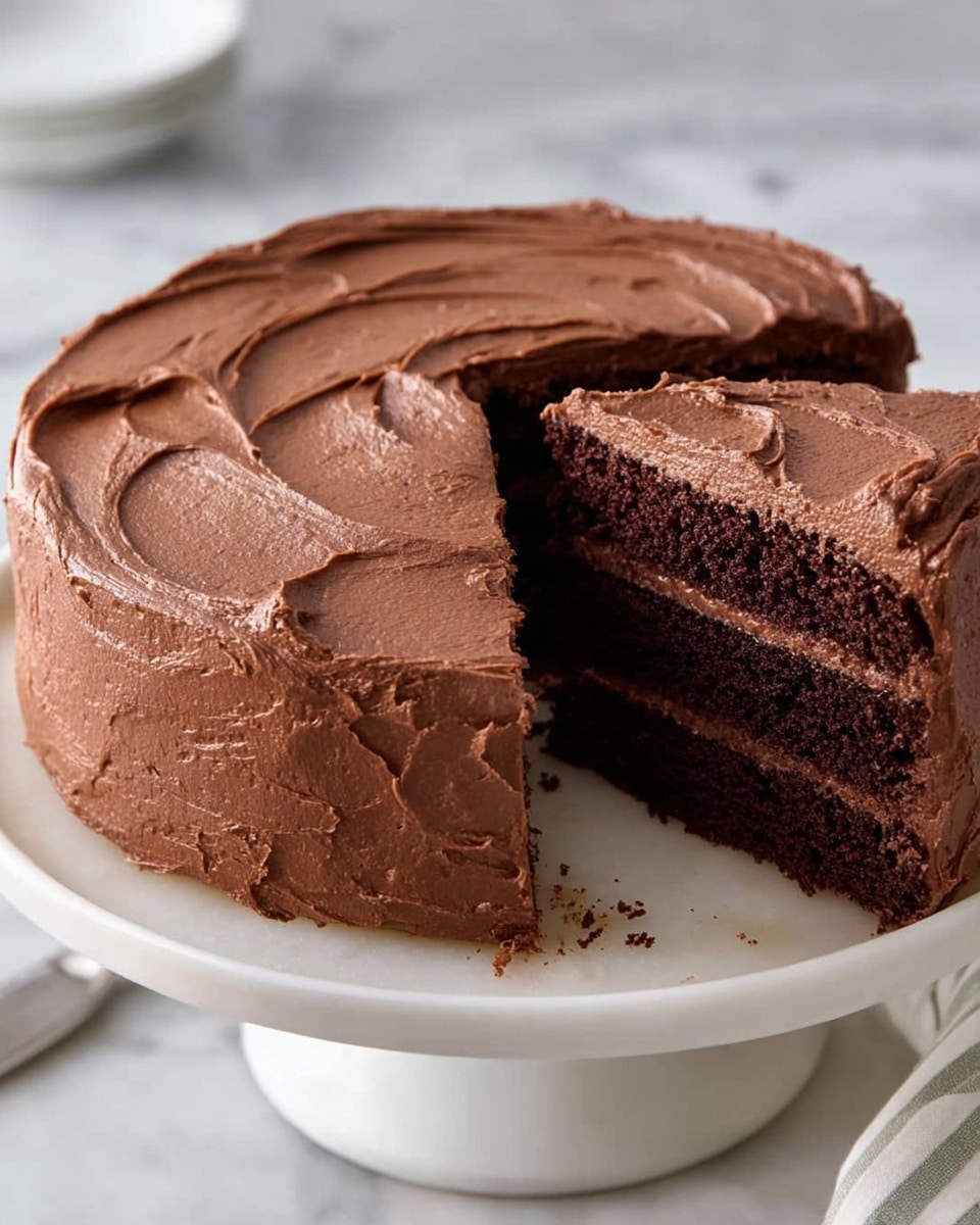 A round chocolate cake with two thick dark brown soft and moist layers is shown on a white cake stand. Between the layers and covering the entire cake is a smooth, rich chocolate frosting with textured swirls on the top and sides. A wedge-shaped slice is cut out and slightly pulled from the main cake, revealing the even, dense layers inside. The background is a white marbled texture. photo taken with an iphone --ar 4:5 --v 7