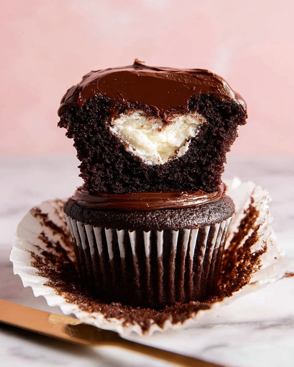 The image shows a rich, dark chocolate cupcake split into two parts, layered one on top of the other on an open white paper liner. The bottom layer is a dense, moist chocolate cake with smooth, glossy chocolate frosting. The top half reveals the inside of the cupcake, showing a thick layer of soft white cream filling in the center, surrounded by dark chocolate cake and topped again with a thick, shiny chocolate frosting that looks slightly textured. The cupcake sits on a white marbled surface with a gold knife nearby that has some chocolate spread on it, and the background is a soft pink gradient. photo taken with an iphone --ar 4:5 --v 7